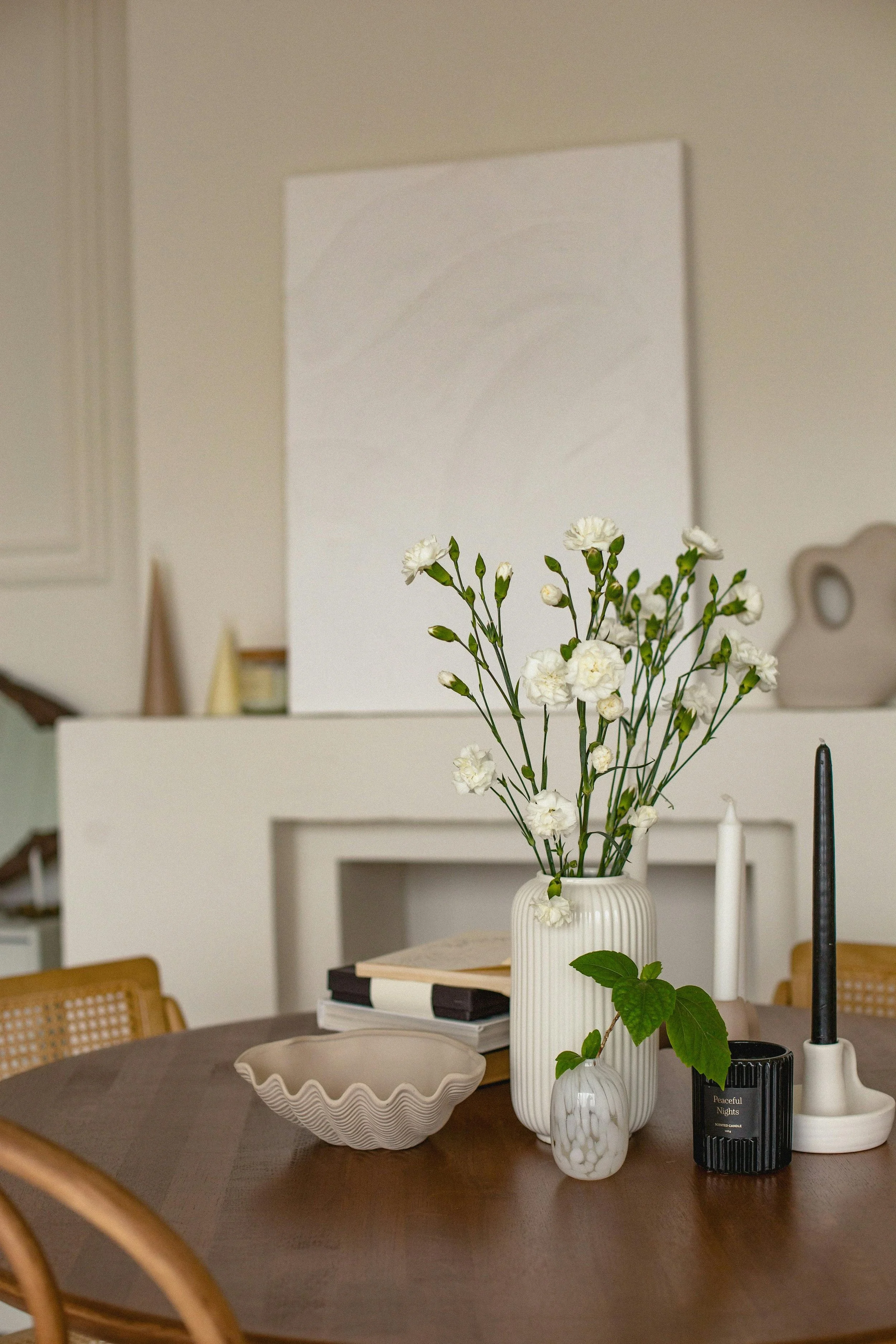 A dining table with a beige ceramic bowl, a white striped vase with white flowers, a small white marble-patterned vase with green leaves, a black scented candle, and two white candles on black holders. In the background, there is a white wall with a blank square canvas and some furniture.