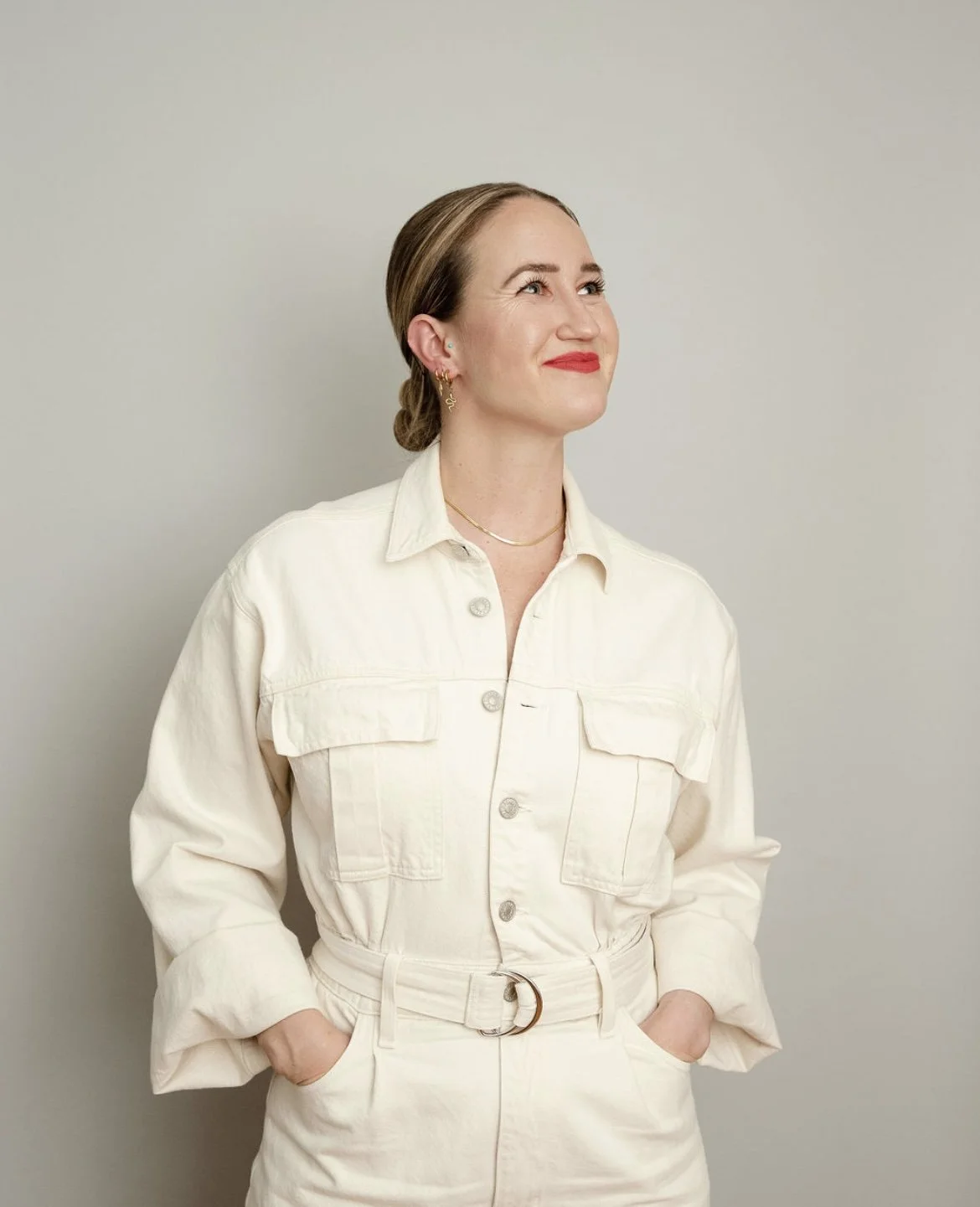 A woman with light brown hair styled in a low bun, wearing a cream-colored button-up jumpsuit with chest pockets, standing against a plain light gray background, looking upwards and smiling.