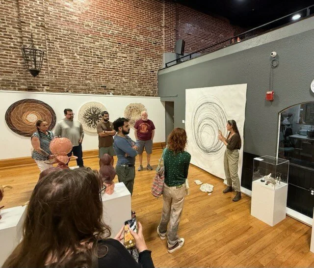 an interior view of the gallery during the artist gallery talk showing a dozen individuals standing among the artwork looking at the artist describe a piece on the wall