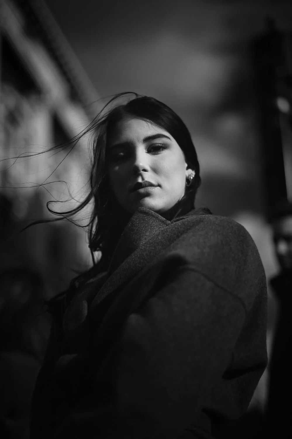 A black-and-white portrait of a young woman with long hair, wearing earrings and a coat, looking at the camera with a serious expression.