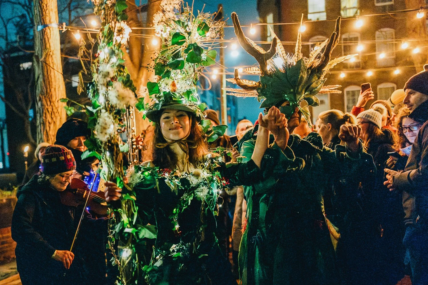 People dressed as forest spirits or nature deities at an outdoor celebration during the evening, with string lights overhead, trees in the background, and a crowd of spectators, some taking photos.