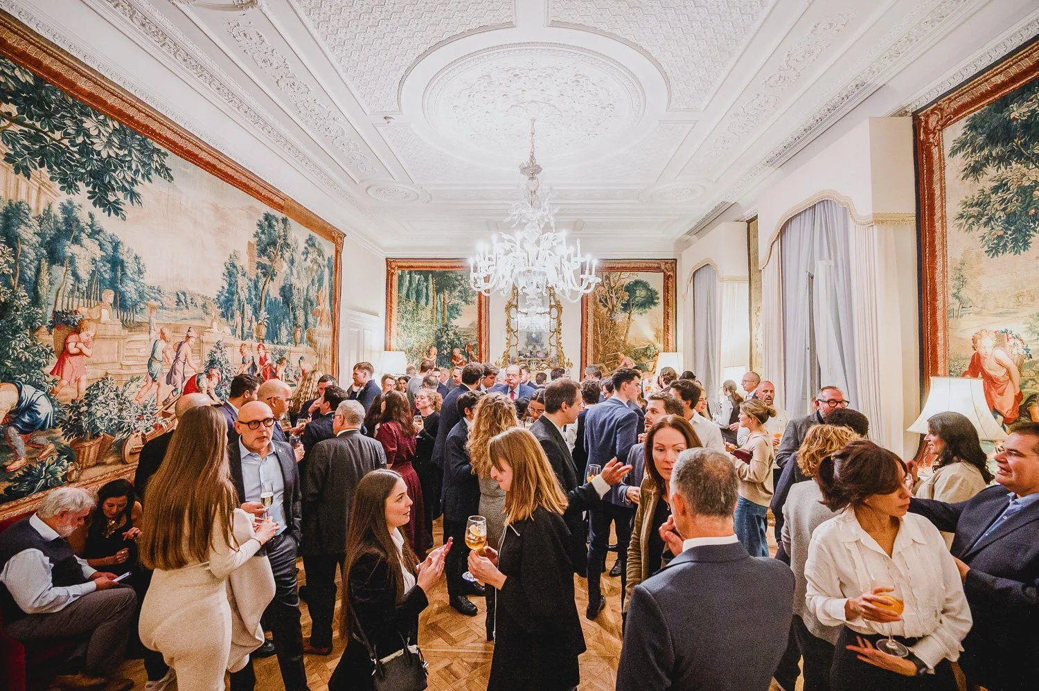A large group of people in formal attire socializing at an elegant indoor event, with ornate walls, large paintings, and a chandelier in the ceiling.
Phocus Collective Event Photography.