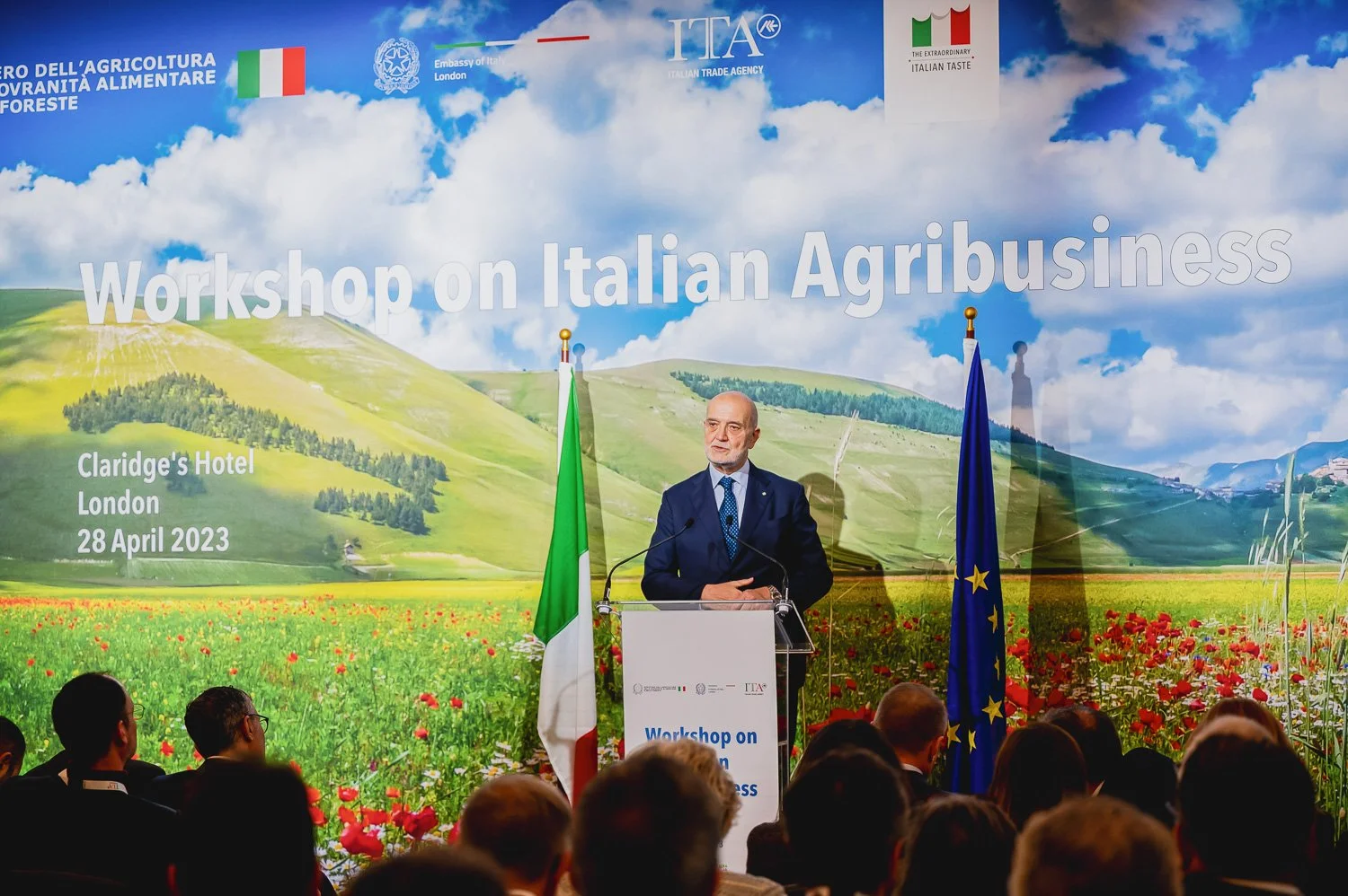 A man speaking at a podium during the workshop on Italian Agribusiness at Claridge's Hotel in London on April 28, 2023, with attendees seated in front. The background features a scenic landscape with rolling green hills and wildflowers, and the backd