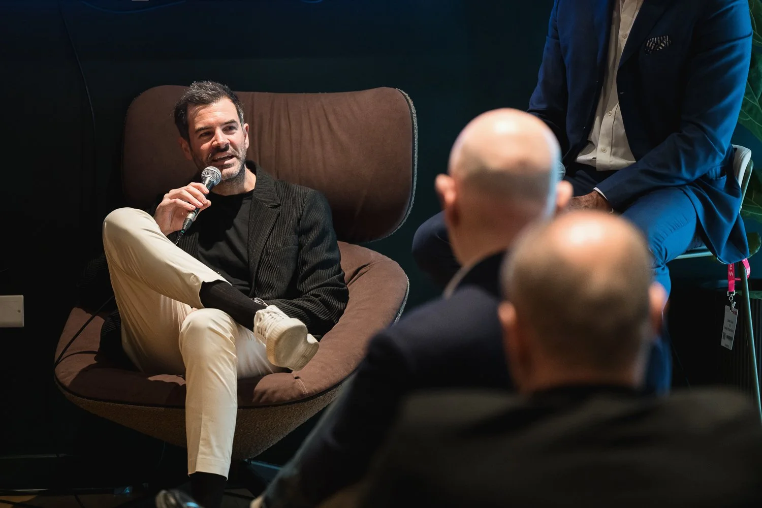 Man in black blazer speaking into a microphone during an indoor panel discussion while seated in a modern chair. Audience members are visible in the foreground.