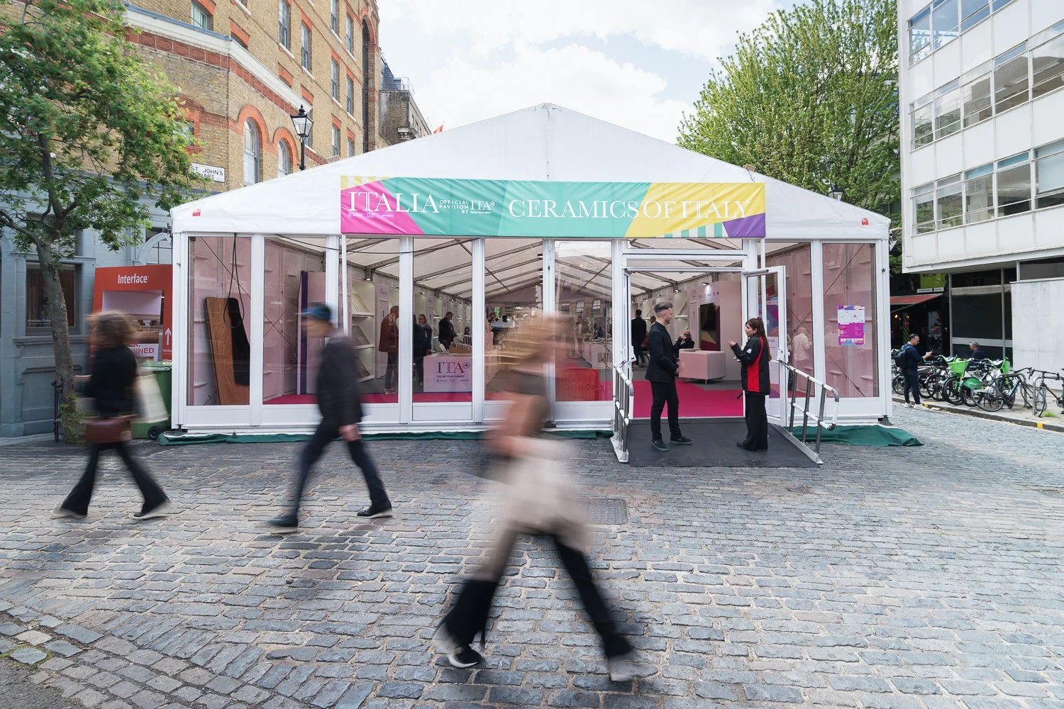 A promotional event tent for ceramics of Italy at the Italy Pavilion, with a rainbow-colored banner, situated outdoors on cobblestone pavement, with people walking by and engaging at the entrance.