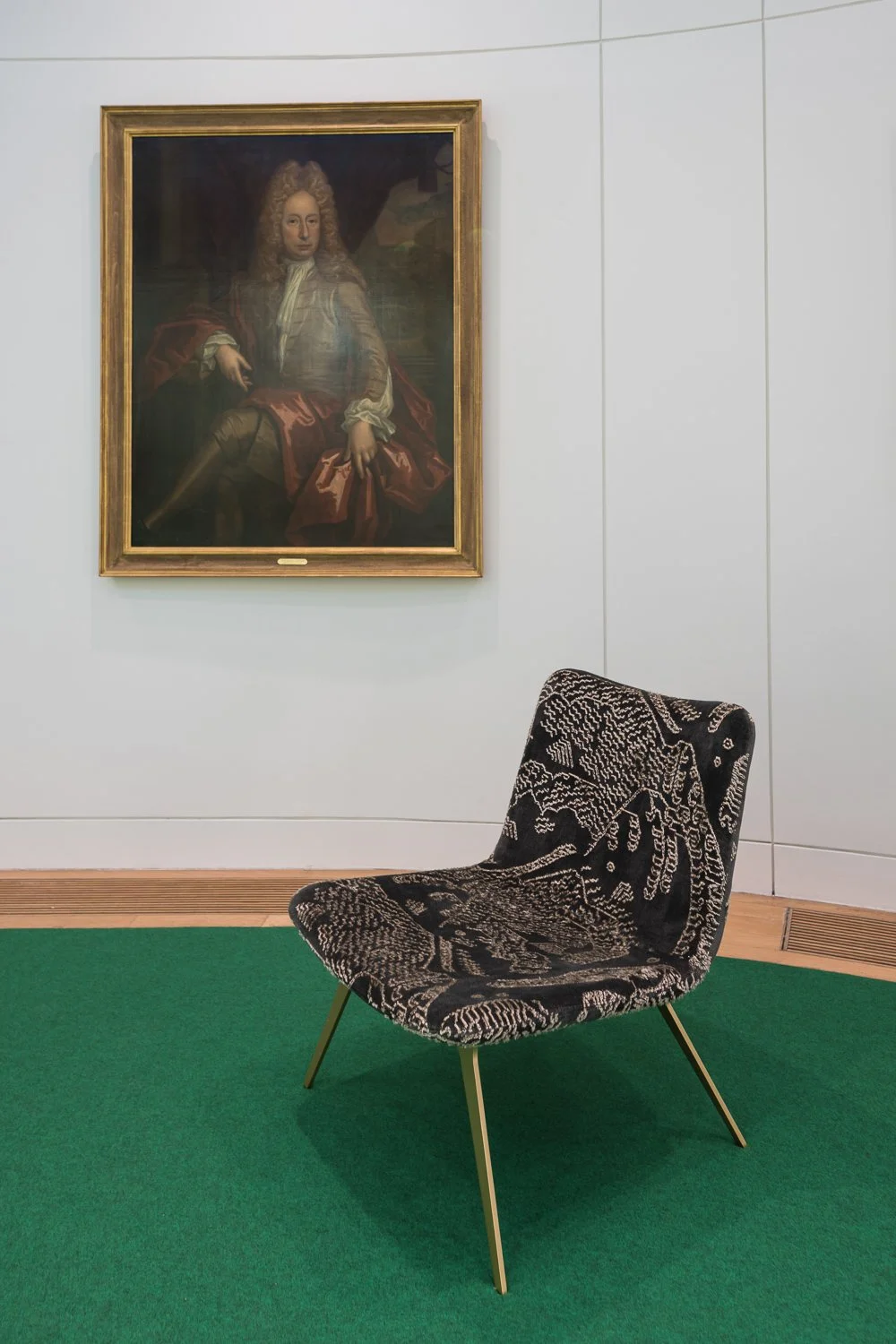 A black and white patterned chair with gold legs, placed on green carpet, in front of a white wall with a portrait painting hanging above.