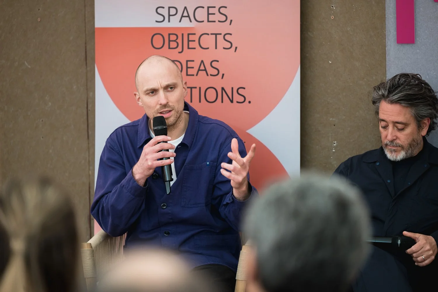 Two men seated at a panel discussion, with one speaking into a microphone and gesturing with his hand, and the other looking down at his notes, in front of a backdrop with the words 'SPACES, OBJECTS, IDEAS, CREATIONS'.