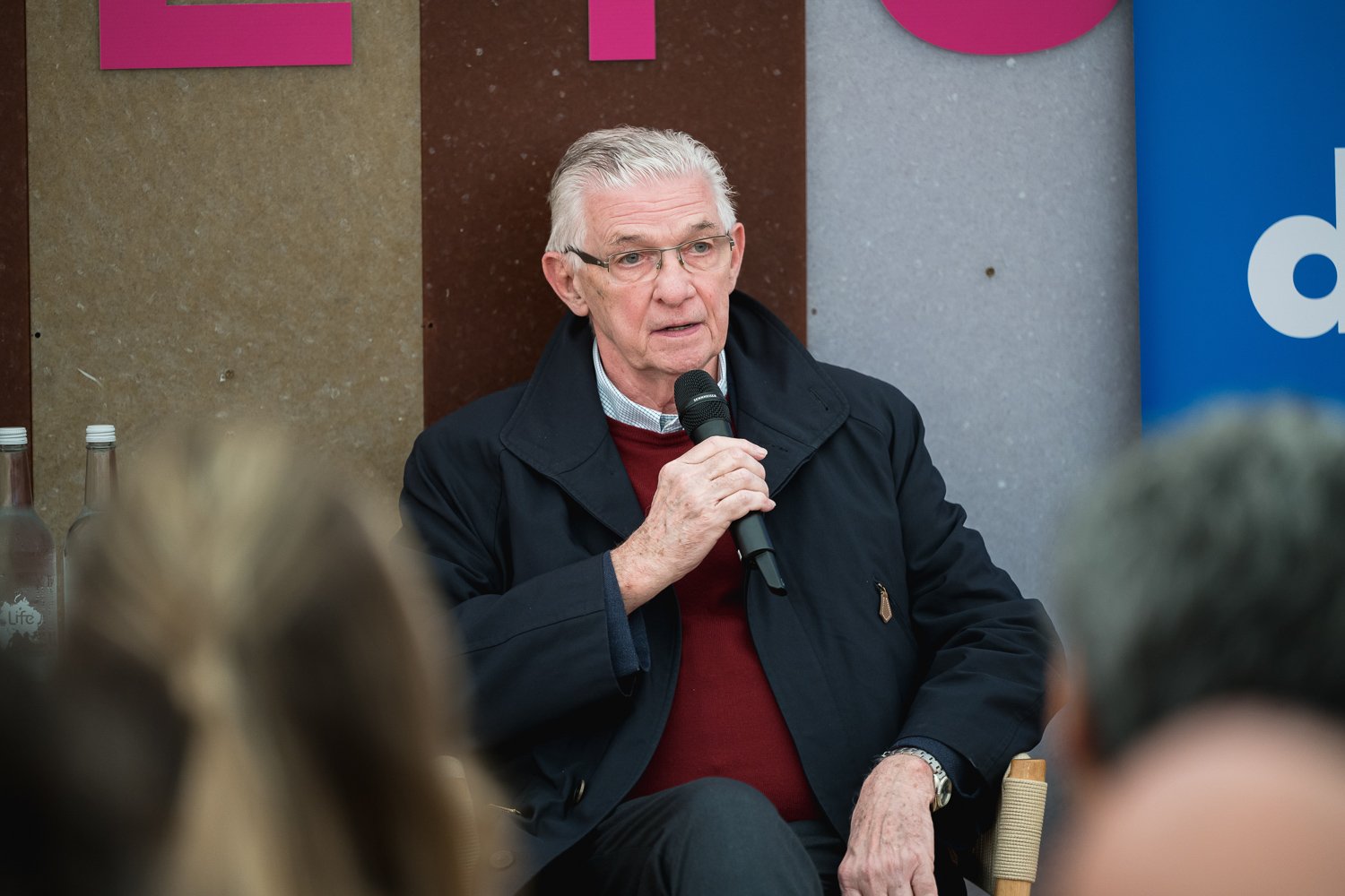 An elderly man with silver hair and glasses speaking into a microphone during a discussion or presentation, seated in a room with colorful wall panels.
