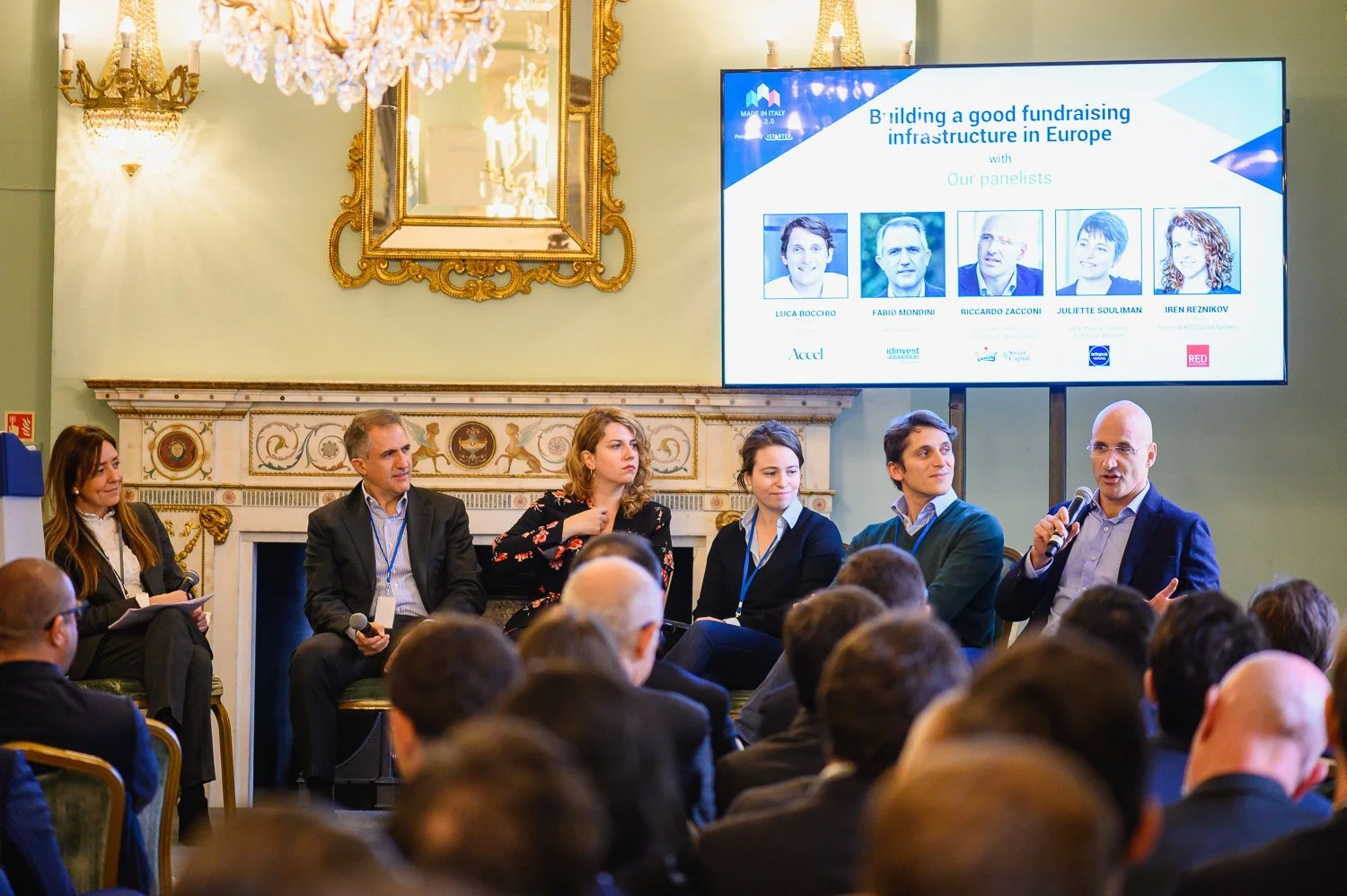 A panel discussion at a conference with six individuals sitting and listening, with one person speaking into a microphone. There is a large screen behind them displaying the event's title, panelists' photos, and names, in a decorated room with chande