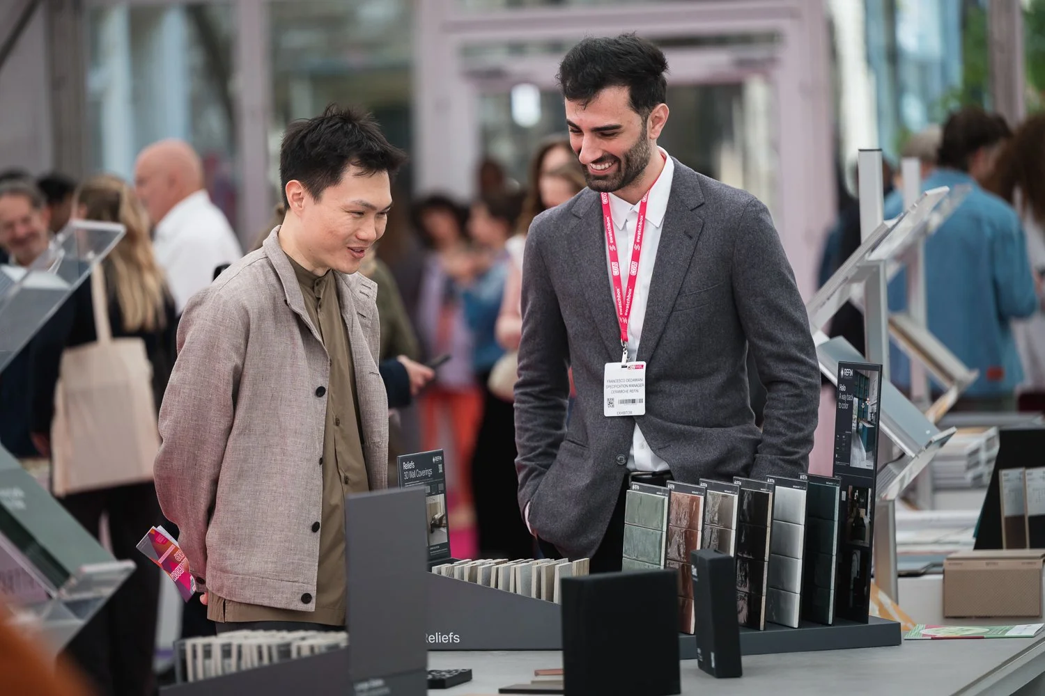 Two men at a trade show booth, looking at various samples of material or surface finishes on display.