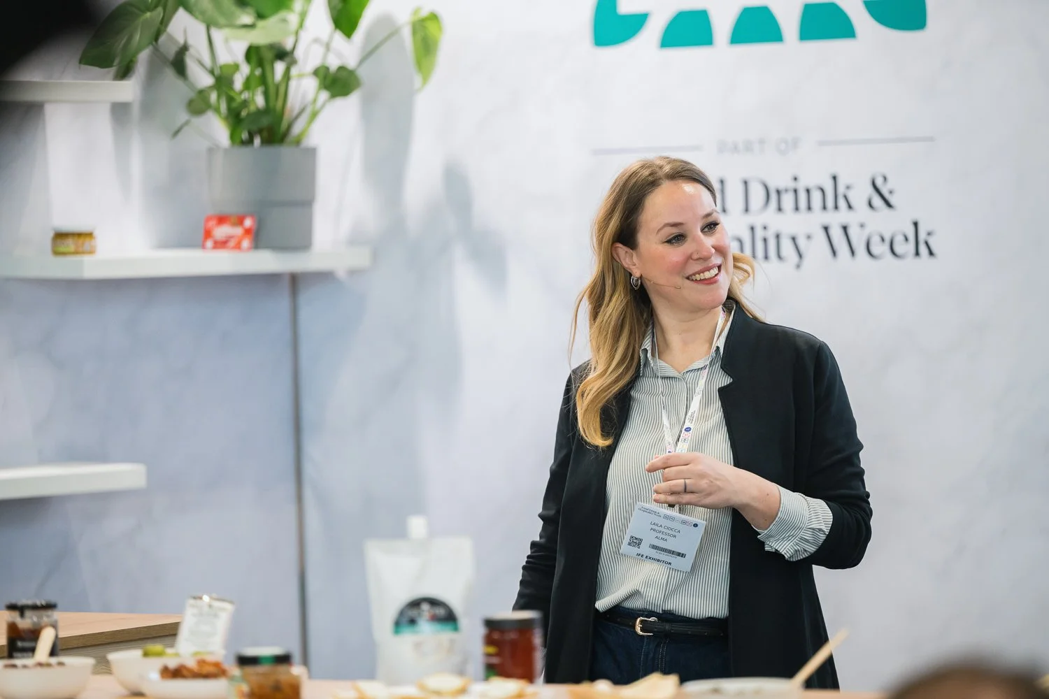 A woman smiling at a food or beverage event with a name badge, standing behind a table with food jars and bowls, and a background sign indicating an food and drink quality week event.