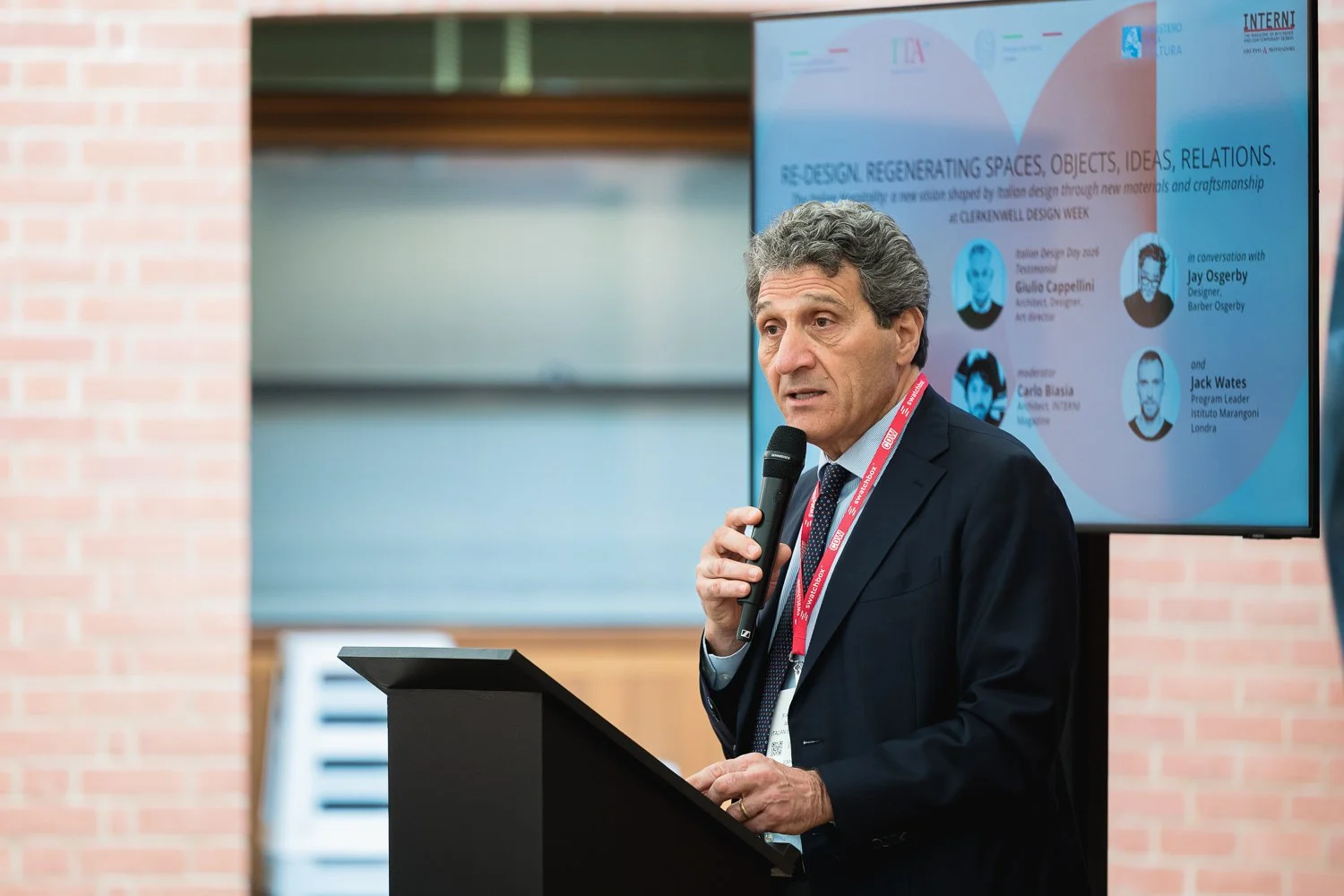A man in a dark suit speaking into a microphone at a podium during a conference or presentation. Behind him is a large screen displaying a presentation slide with text and images of other speakers.