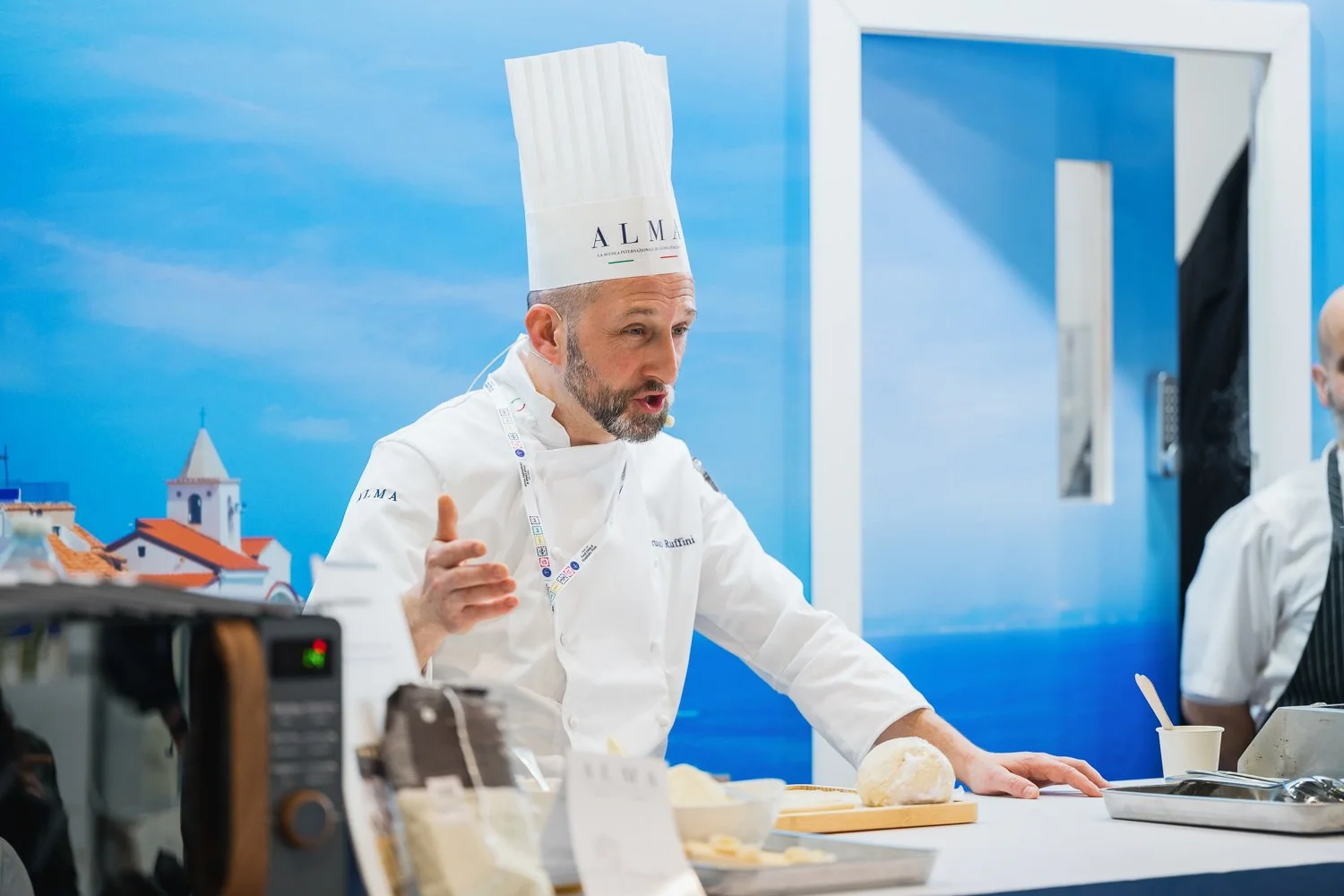A professional chef wearing a tall white chef hat and white uniform is explaining something while preparing food in a kitchen with a blue wall background.
