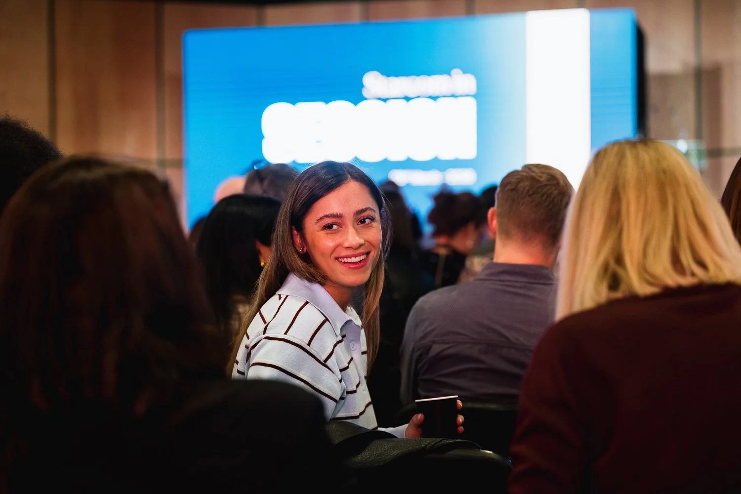 A woman with long brown hair wearing a white striped shirt is smiling and looking at the camera during a conference or presentation, with other attendees seated around her and a large screen in the background.