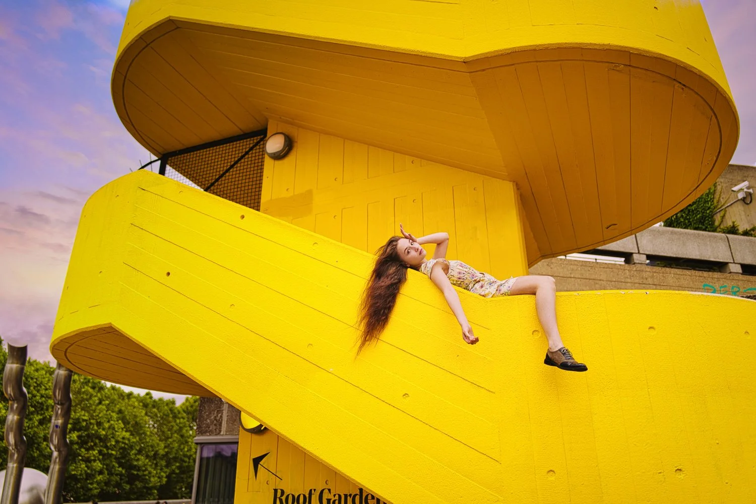 A woman with long hair lying on a yellow, curved architectural structure, with a sunset sky in the background.