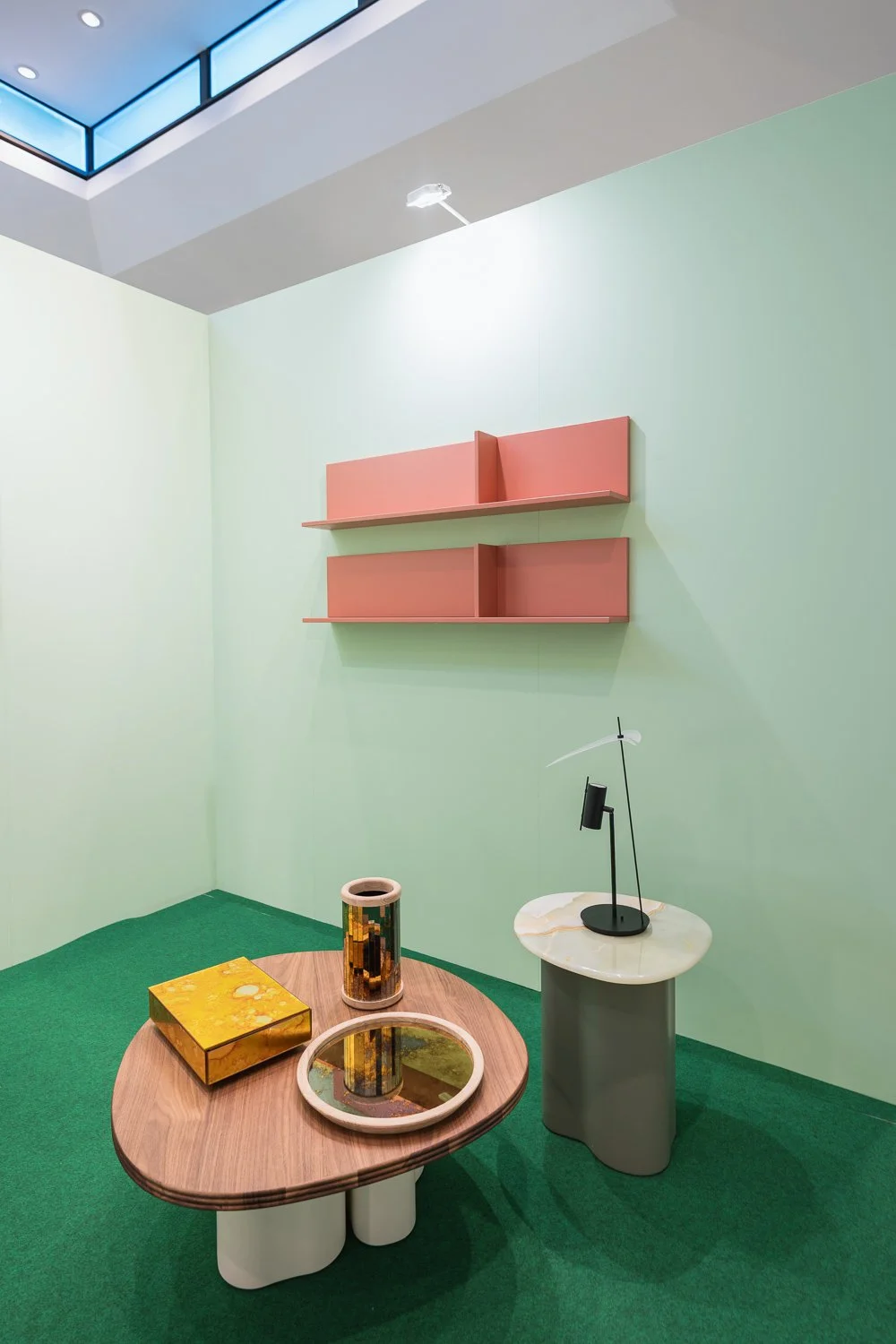 Minimalist interior with light green walls, a green carpeted floor, a wooden table with decorative items, and pink wall-mounted shelves.