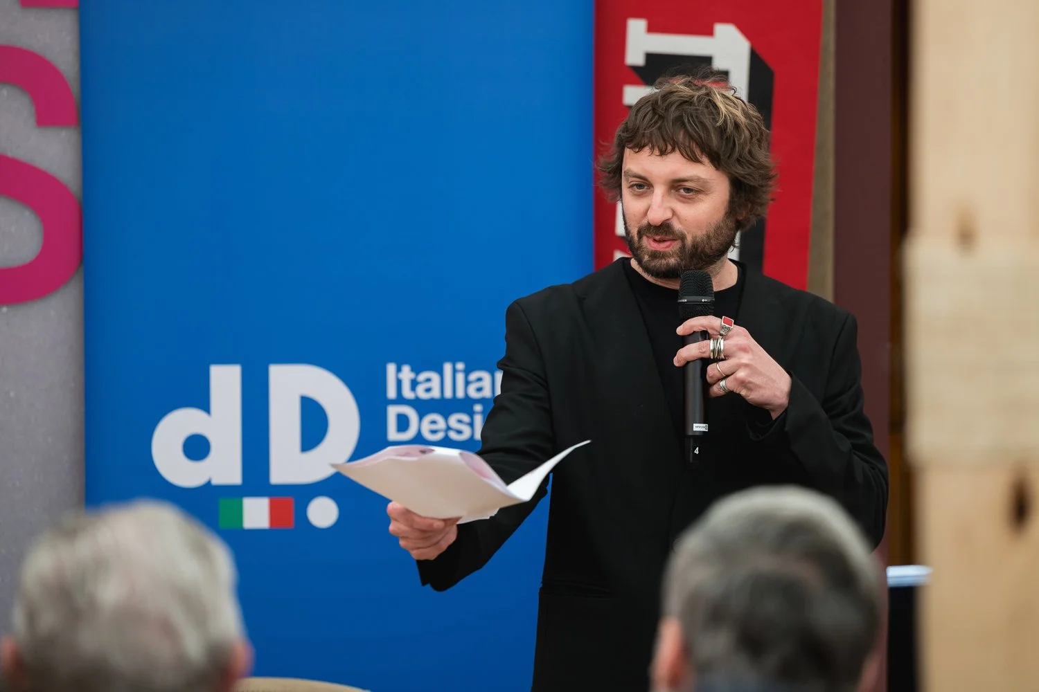 A man with brown curly hair and a beard speaking into a microphone while holding an open paper, standing in front of a blue and red sign with the words 'dD Italian Design'.