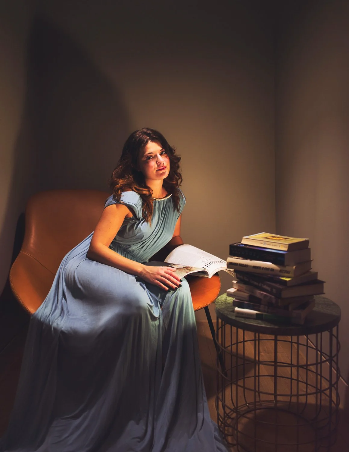 A woman with dark hair in a blue gown sitting on a yellow chair, looking at the camera, with a stack of books on a small round table beside her.
