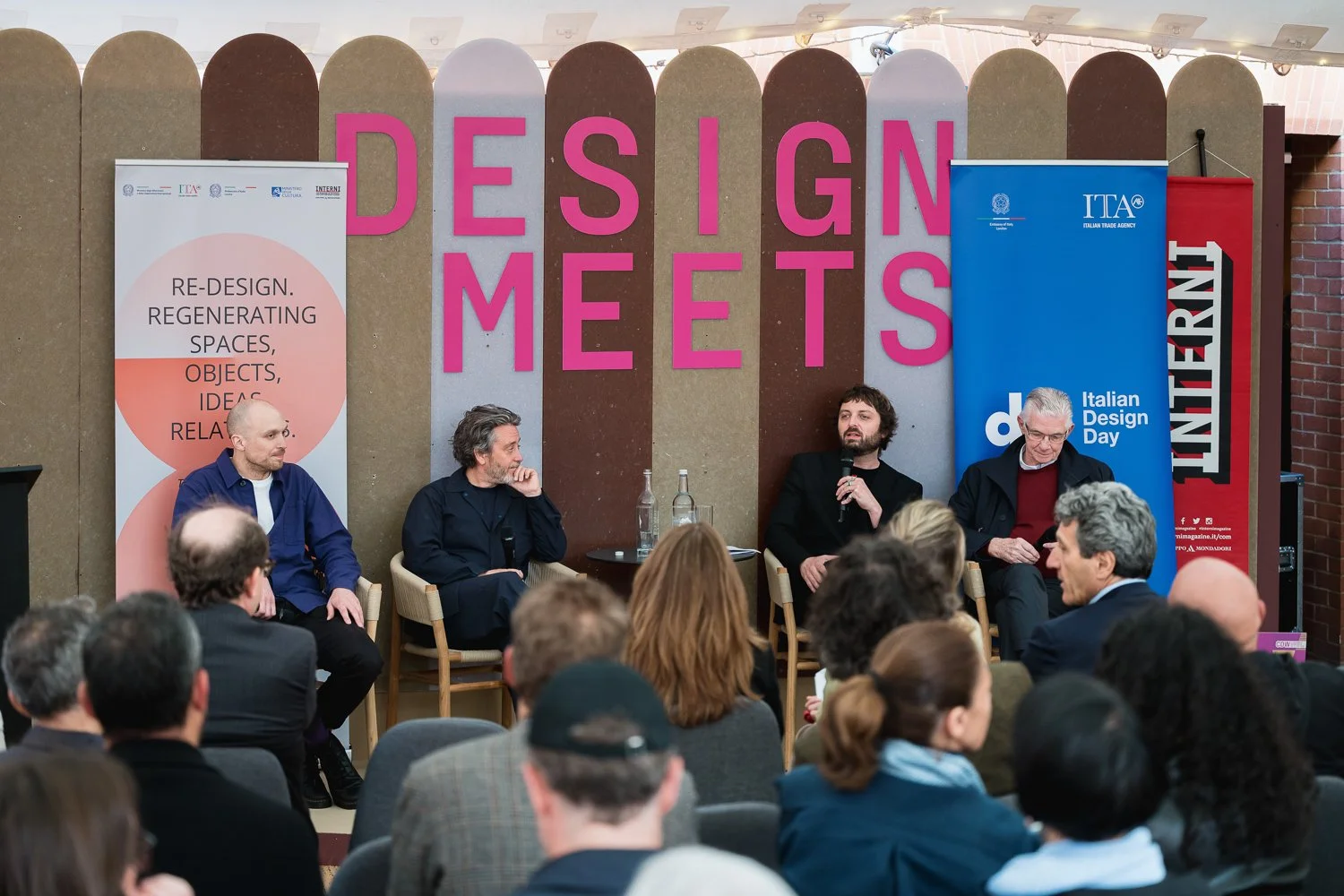 A panel discussion at Italian Design Day with four male speakers seated on stage, one of whom is speaking into a microphone. The stage backdrop has large pink letters spelling 'DESIGN MEETS' and banners from the Italian Trade Agency and related organ