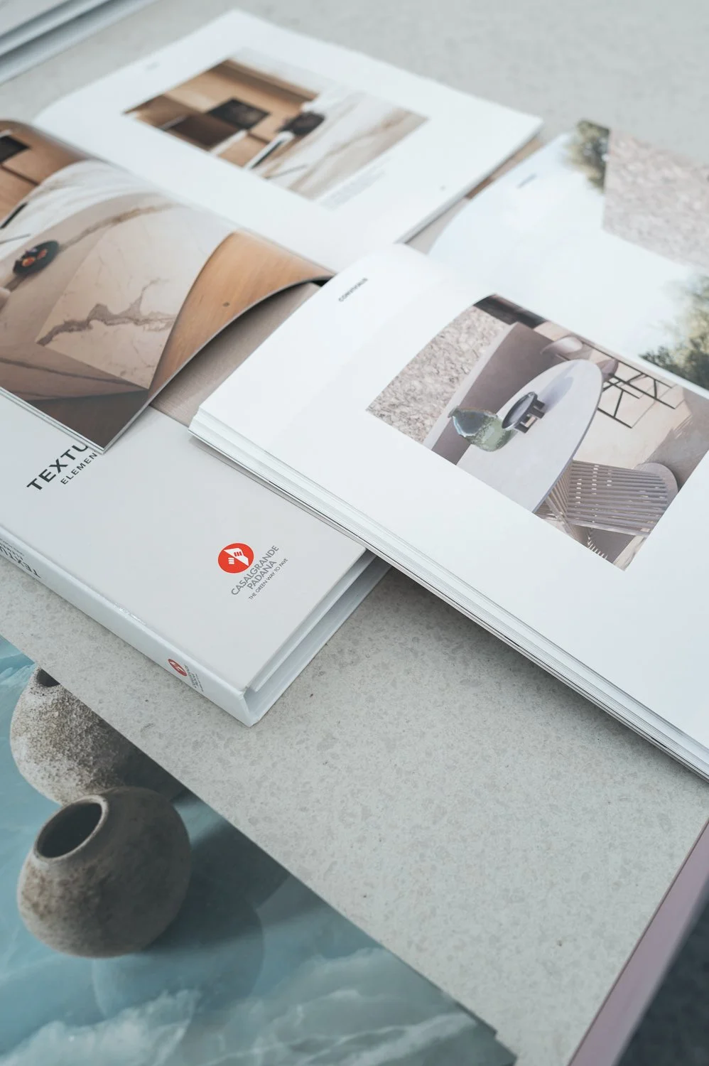 Open design catalog displaying interior decor and furniture images on a light-colored table with a small textured pottery vase nearby.