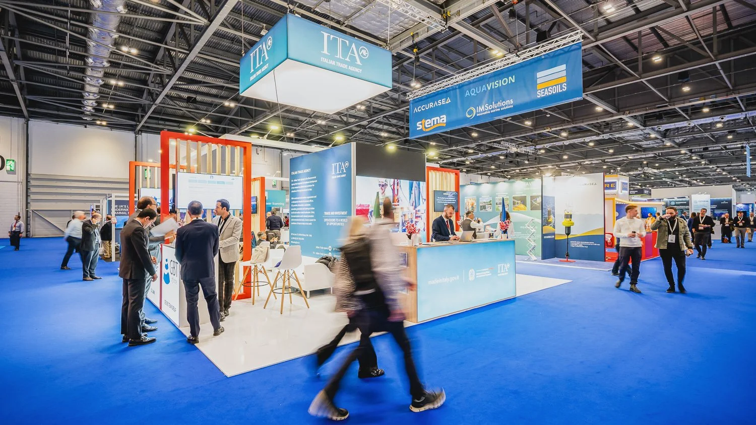 Trade show booth with signs for ITA and other companies, people walking and talking in the exhibition hall with a blue carpeted floor and high ceiling. Phocus Collective Event Photography.