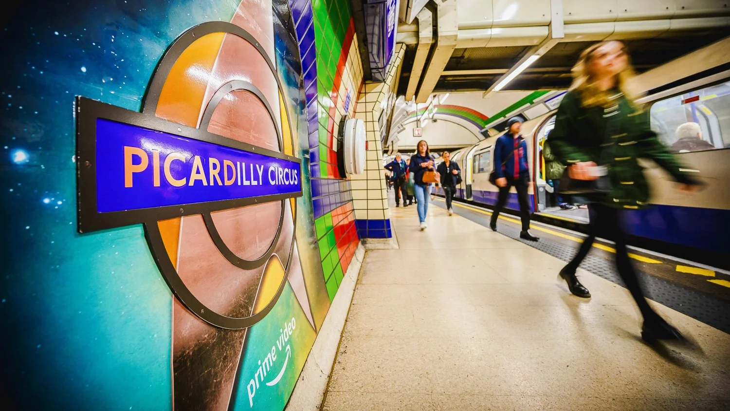 Underground subway station with pedestrians walking along the platform, colorful murals on the walls, and a train arriving at the station. There is a sign for Piccadilly Circus.