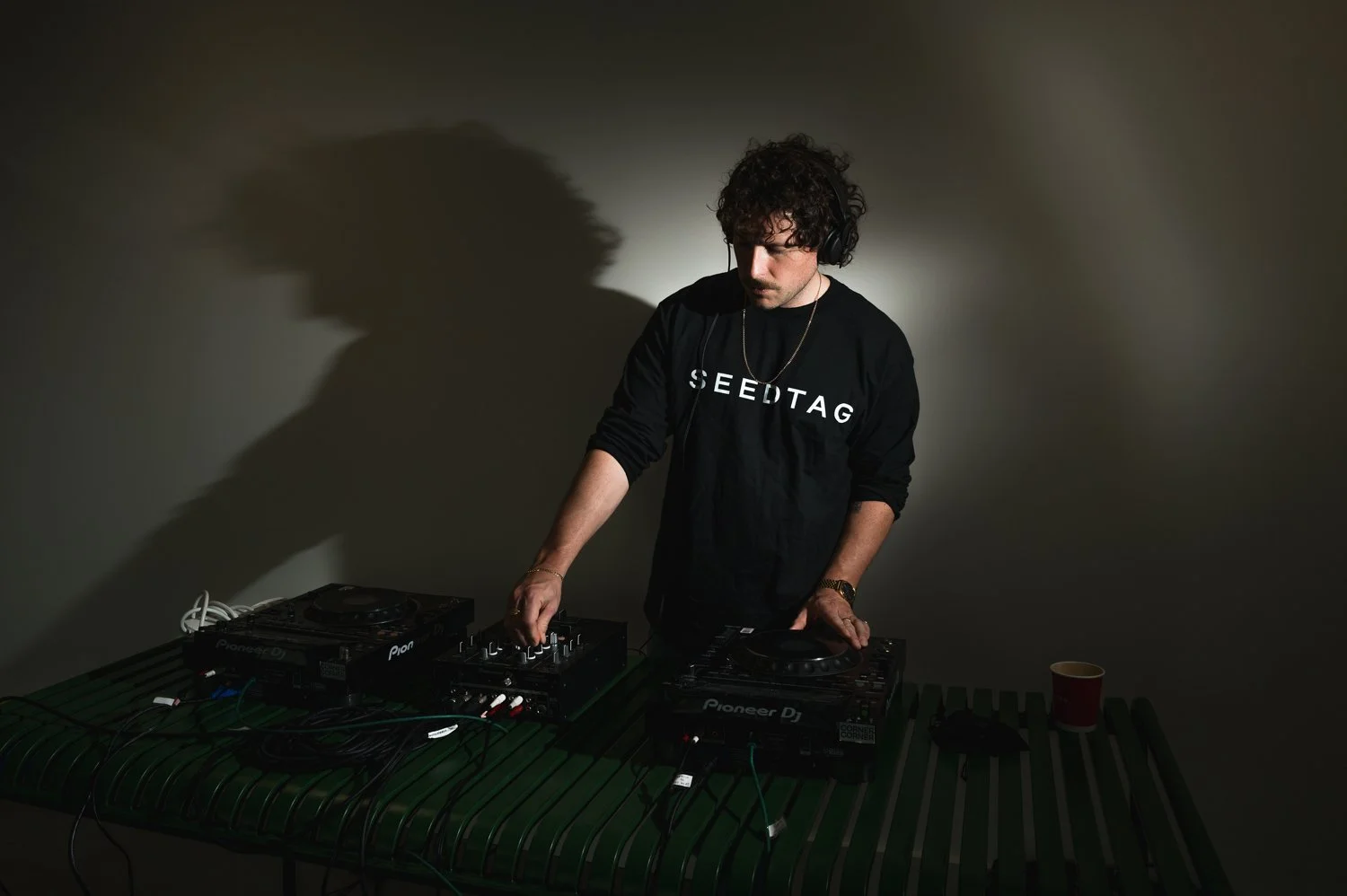 A DJ with curly dark hair, wearing a black sweatshirt with 'SEEDTAG' written on it, is performing. He is using turntables on a green table, with a dark background and dim lighting.