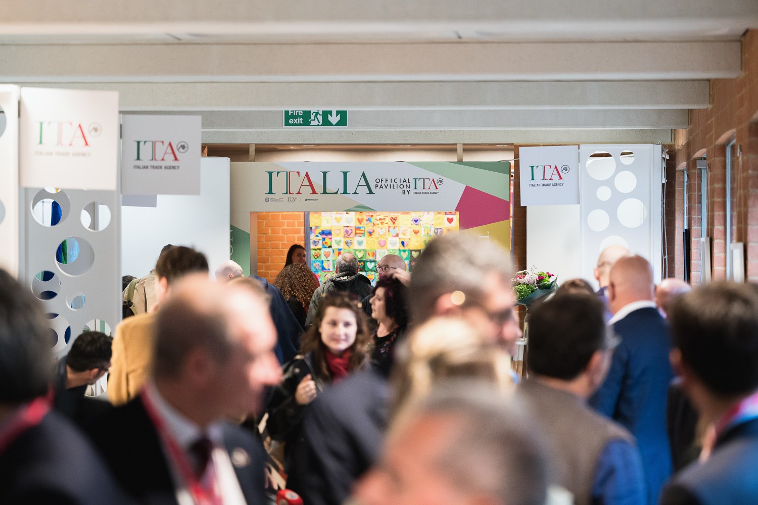 Crowd of people at an Italiana trade agency event with a sign overhead reading 'ITALIA of official pavilion' and colorful artwork in the background.