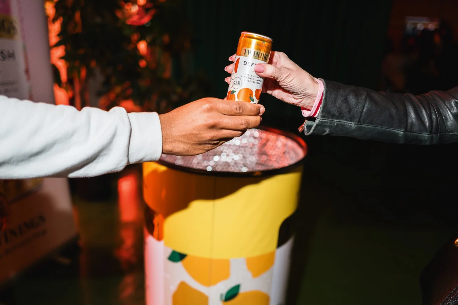 Two people exchanging a can of orange drink over a recycling bin in a dimly lit outdoor setting.