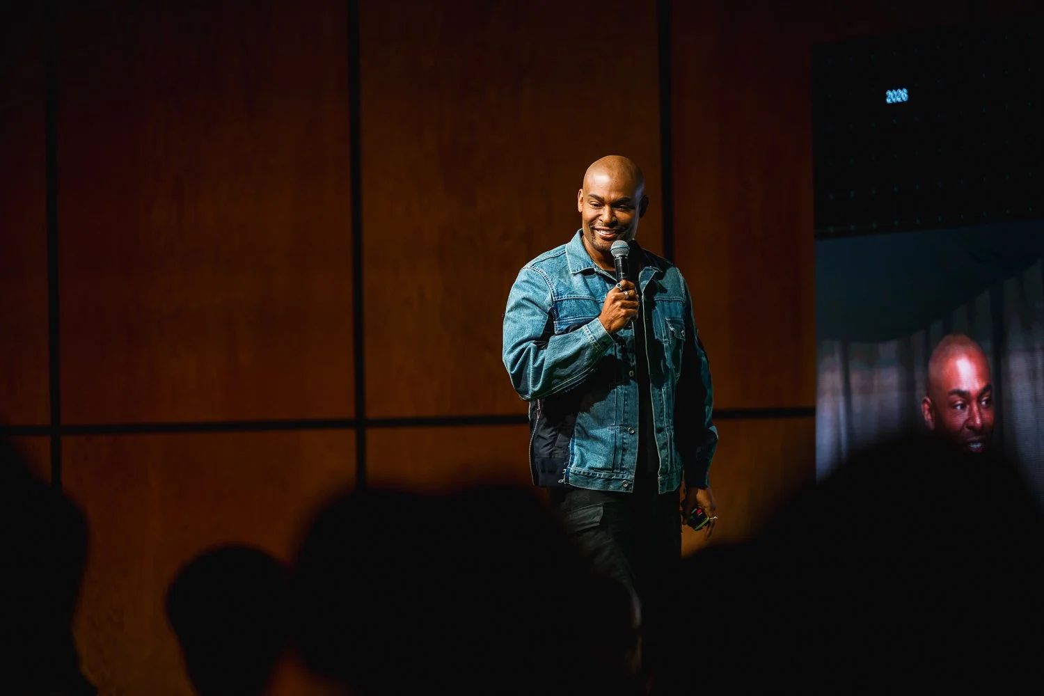 A man with a shaved head wearing a denim jacket stands on a stage, holding a microphone and smiling, with a wooden wall behind him and an audience silhouette in the foreground.