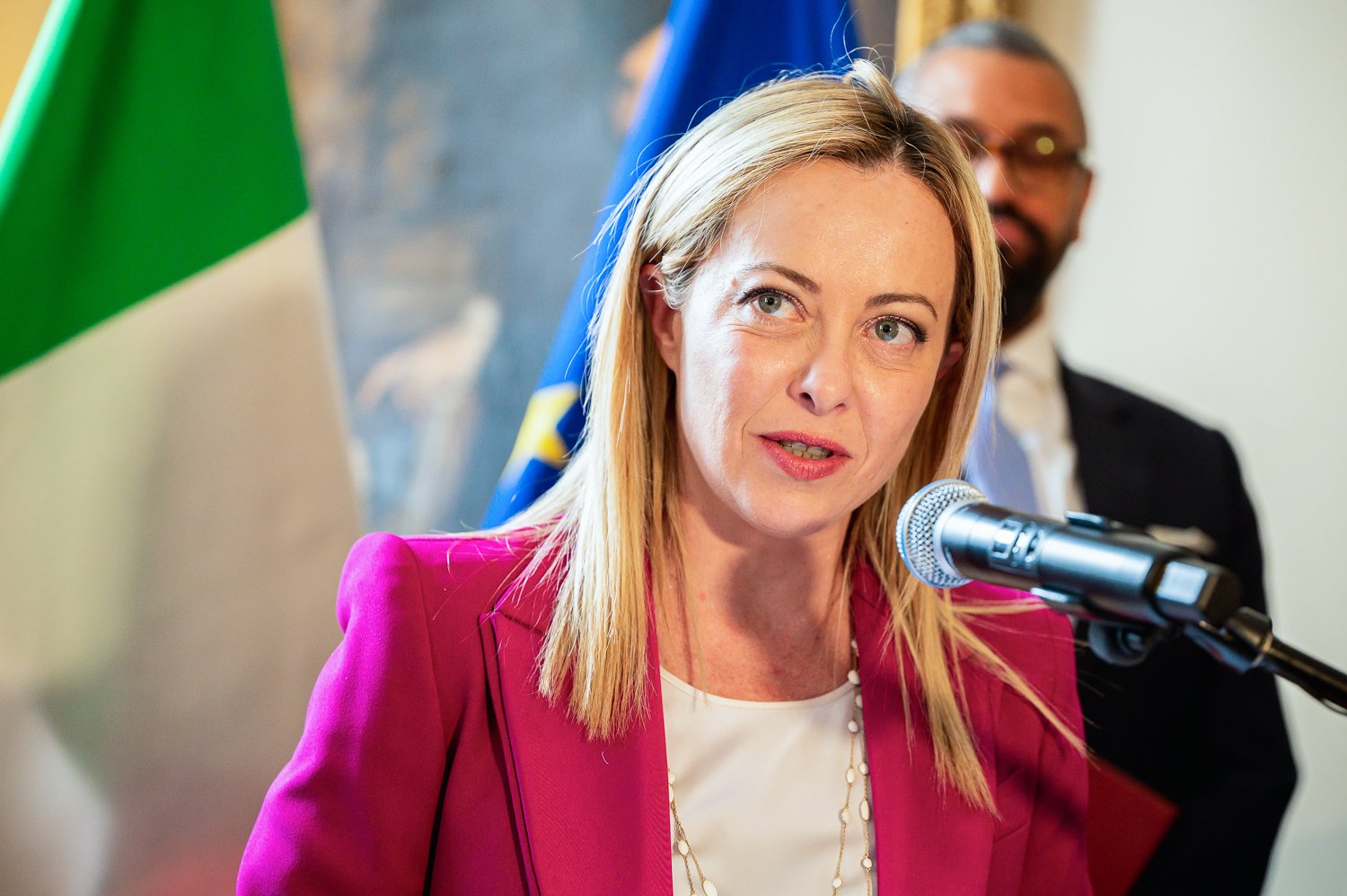 A woman with blonde hair speaking into a microphone during a press conference, with a man in the background and flags behind them.