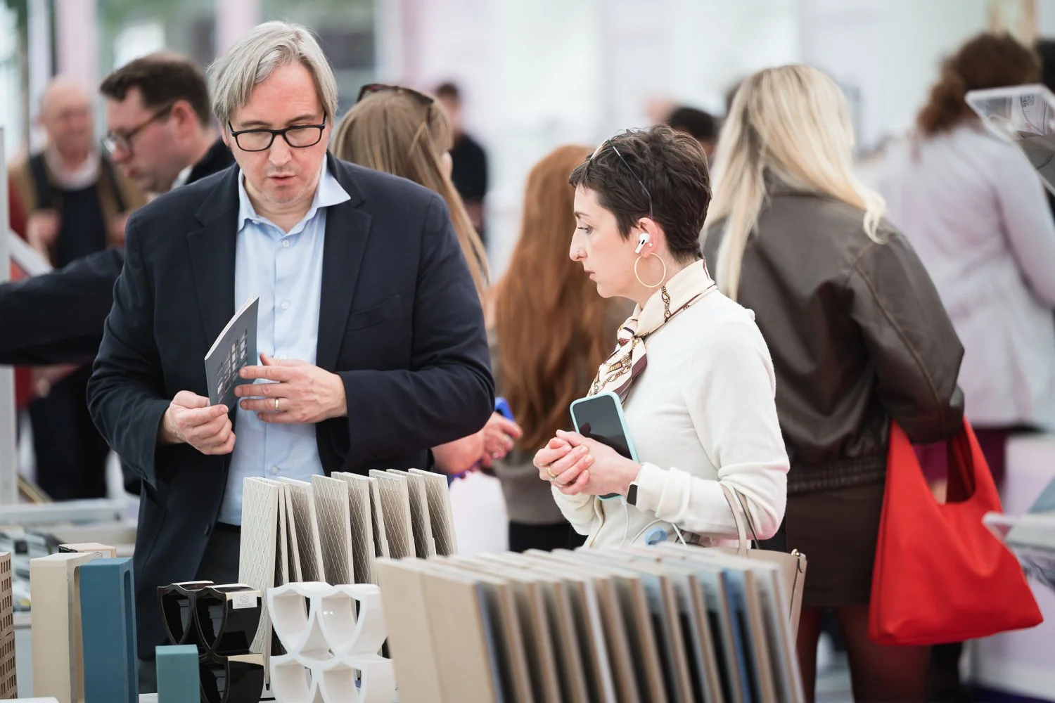Two people browsing different tile samples at an indoor market or store, surrounded by other shoppers.