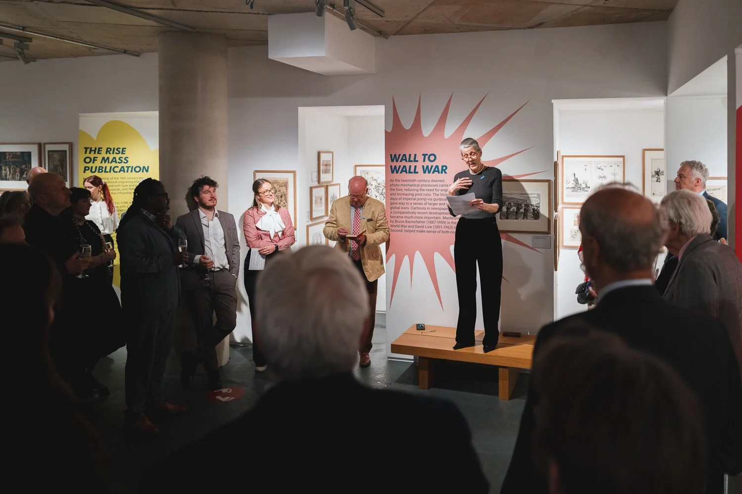A woman stands on a small wooden stage, reading from a paper, in front of an audience at an art gallery. The audience, diverse in age and style, stands and listens. The gallery wall features framed artworks and informational panels, including one tit