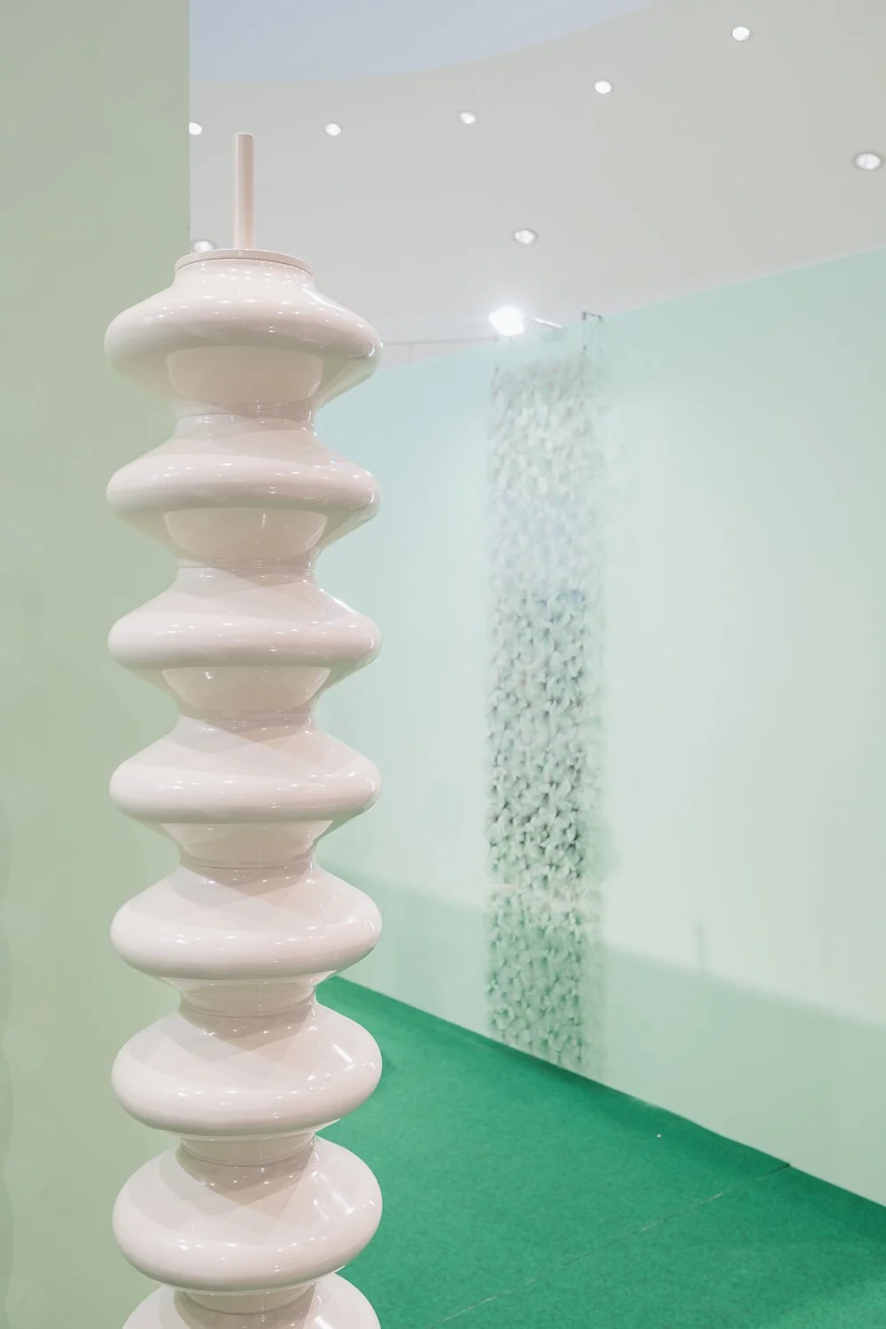 A tall, white, ceramic sculpture with a series of rounded, symmetrical shapes stacked vertically. The sculpture is positioned in front of a mint green wall with a textured vertical art piece and a green carpeted floor.