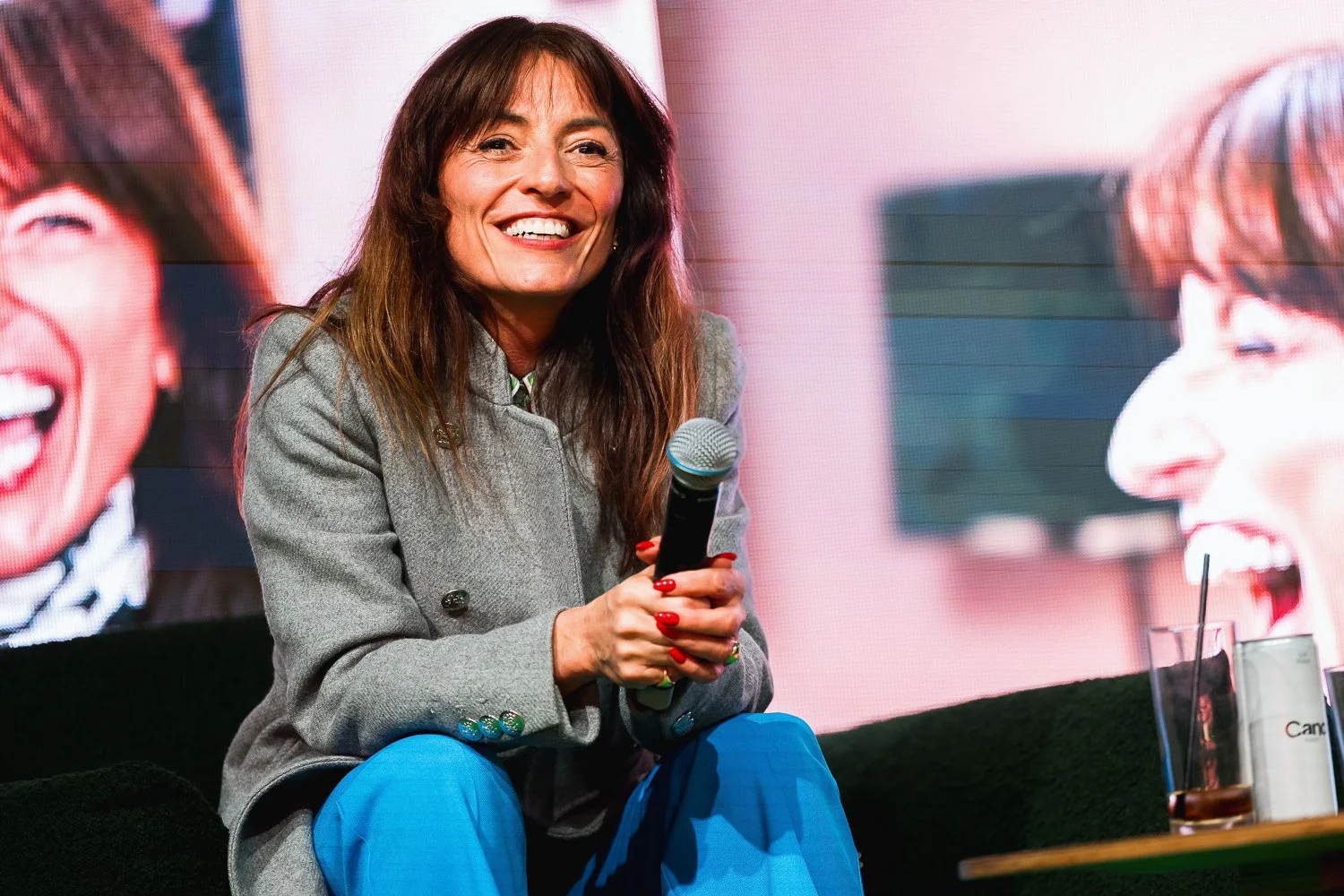 A woman with long brown hair, smiling and holding a microphone, sitting on a black couch in front of a screen displaying her large photo laughing.