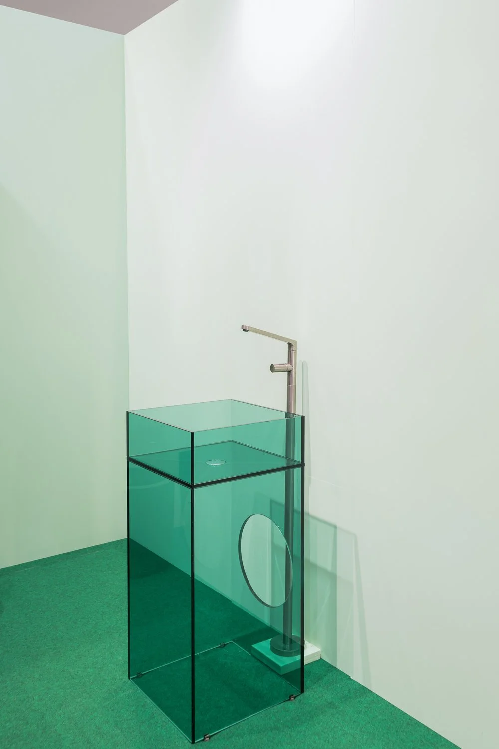 A minimalist room with a white wall and green carpet, featuring a modern glass sink with a metal faucet and a white base, standing behind a transparent glass partition.