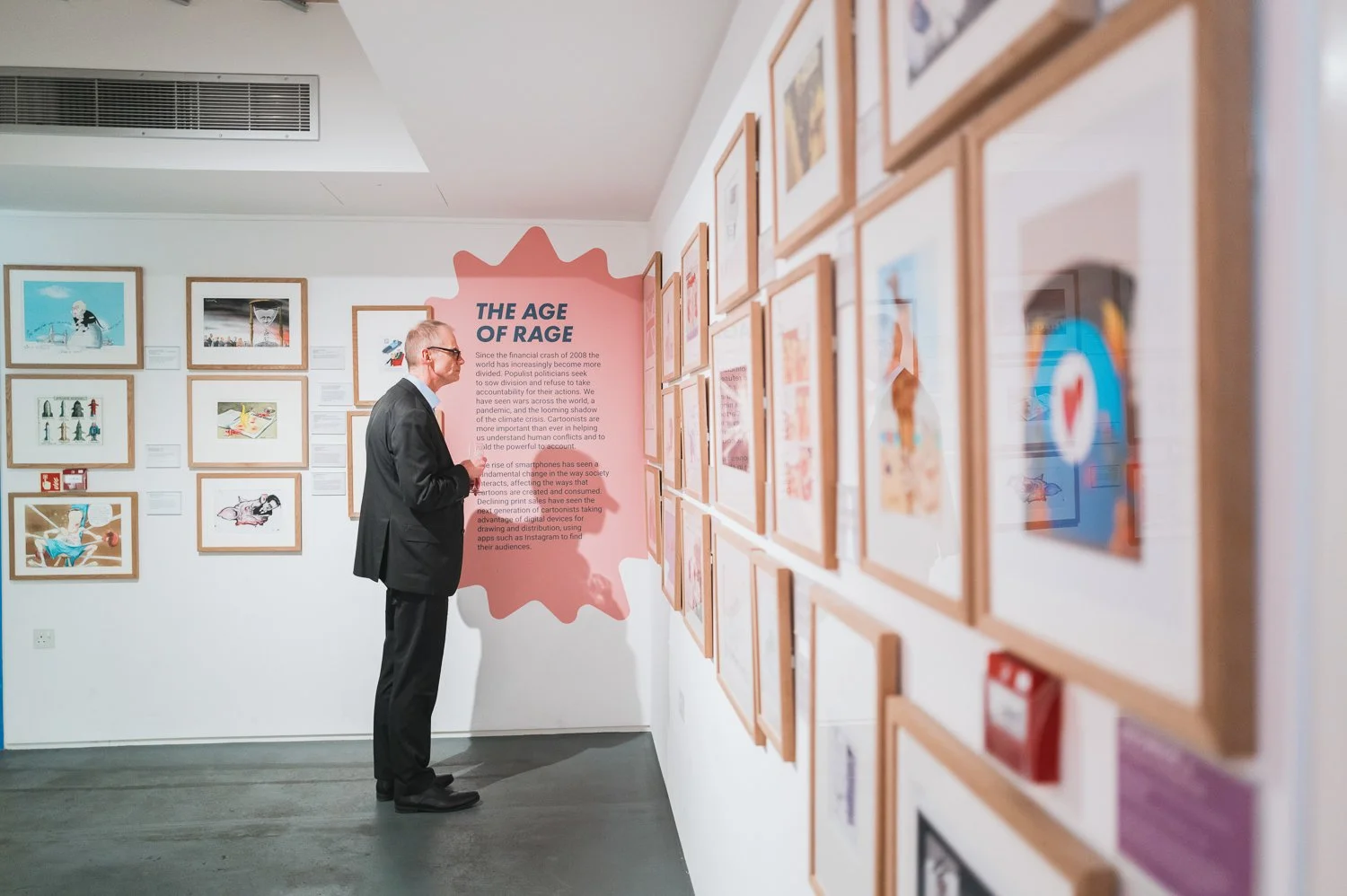 A man in a black suit and glasses looking at framed artwork on a white gallery wall with a pink informational sign titled 'The Age of Rage'.