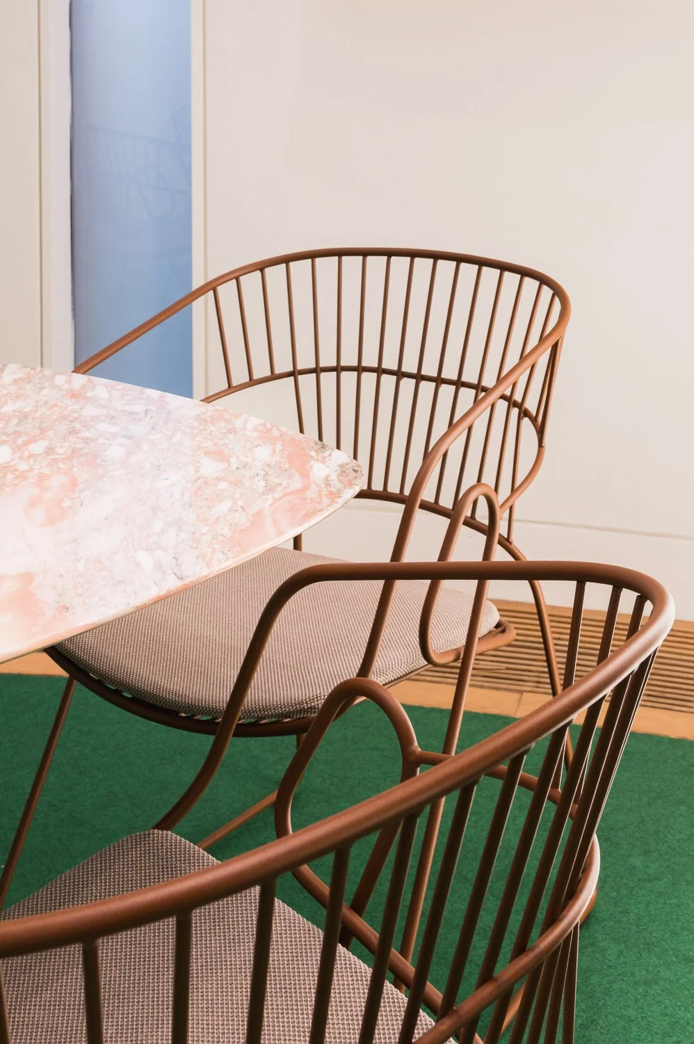 A dining area featuring a pink marbled tabletop, brown metal chairs with beige cushions, on a green carpet with a white wall and a blue door in the background.