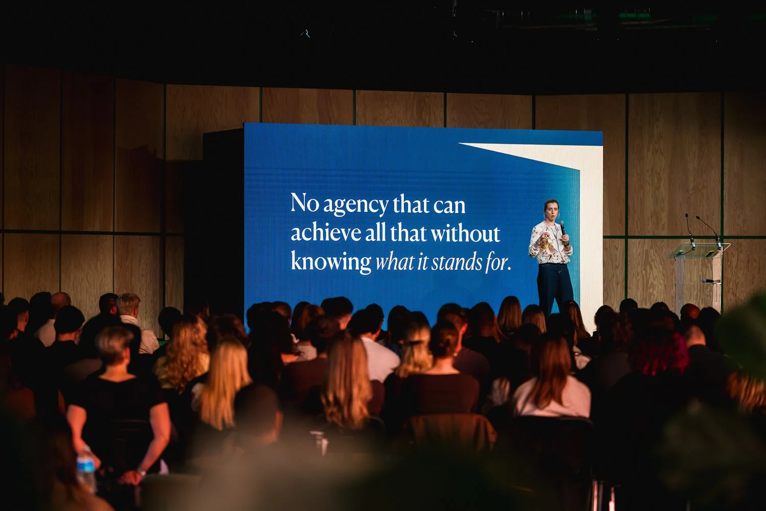 A woman is giving a presentation on stage, holding a microphone, with a large screen behind her displaying a quote about the importance of understanding what an agency stands for. The audience is seated in front of her in a conference room.