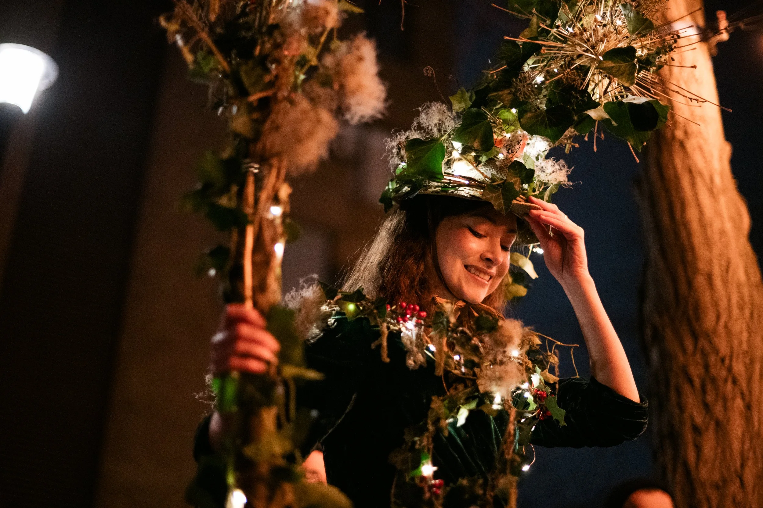 A young woman with long brown hair dressed in a dark outfit, smiling, wearing a festive hat decorated with ivy, berries, and fairy lights, against a dark background.