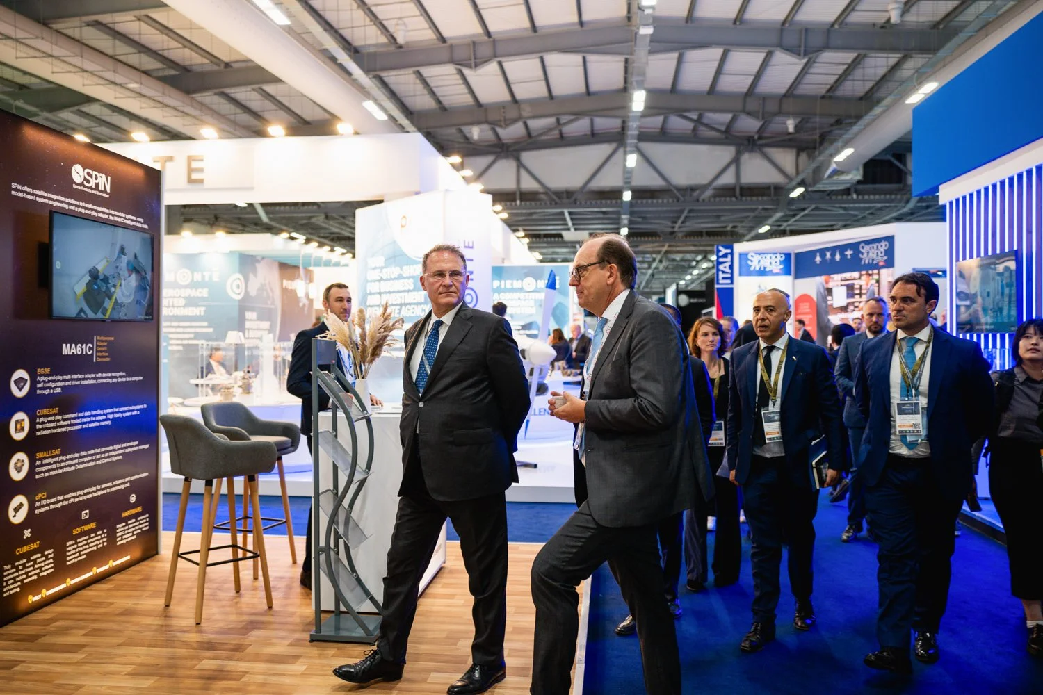 Business professionals and attendees walking and talking at a trade show or conference in an exhibition hall with various booths and displays.