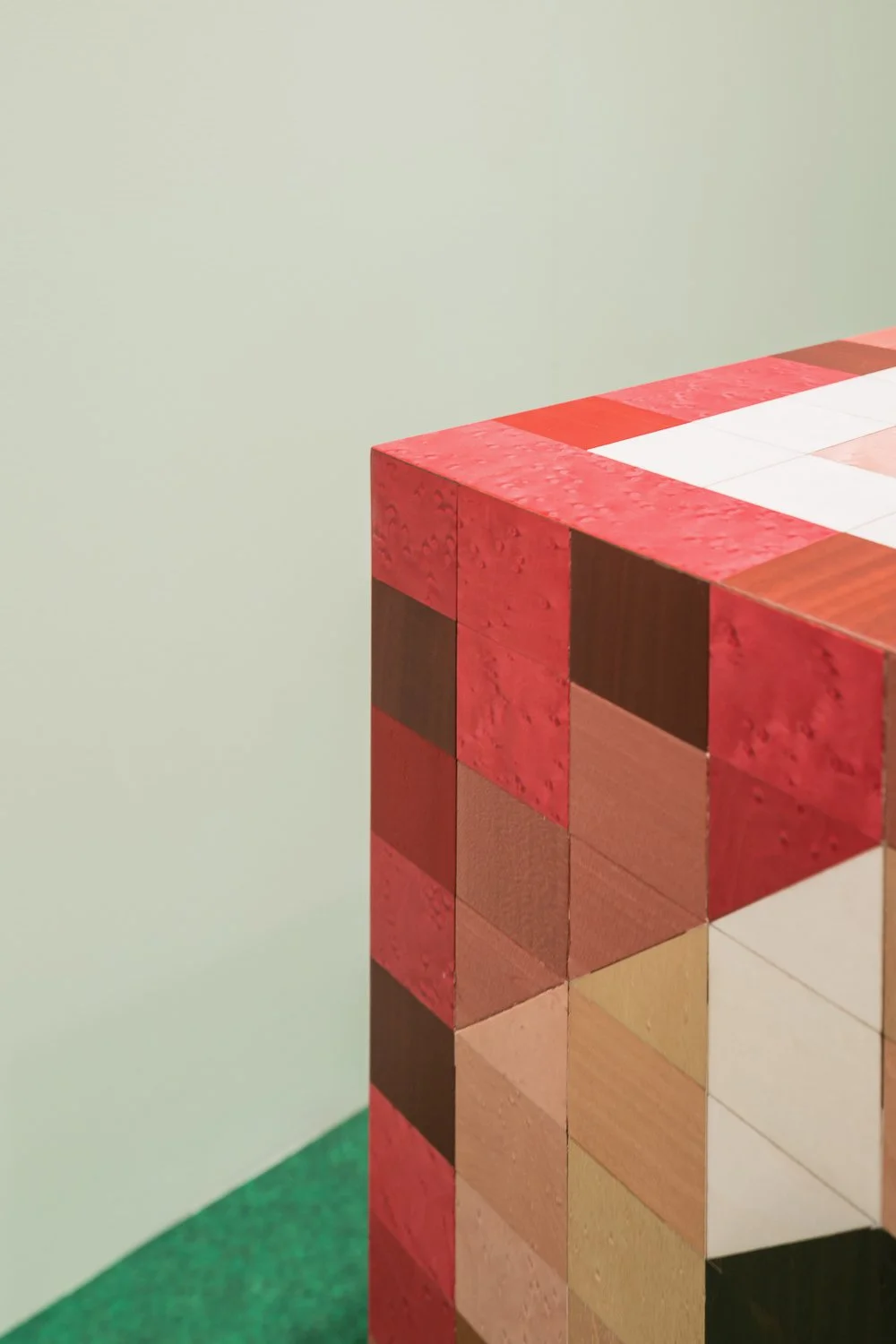 Close-up of a wooden cube structure with red, brown, and beige tiles, positioned in a corner with a green carpet and plain light-colored wall.