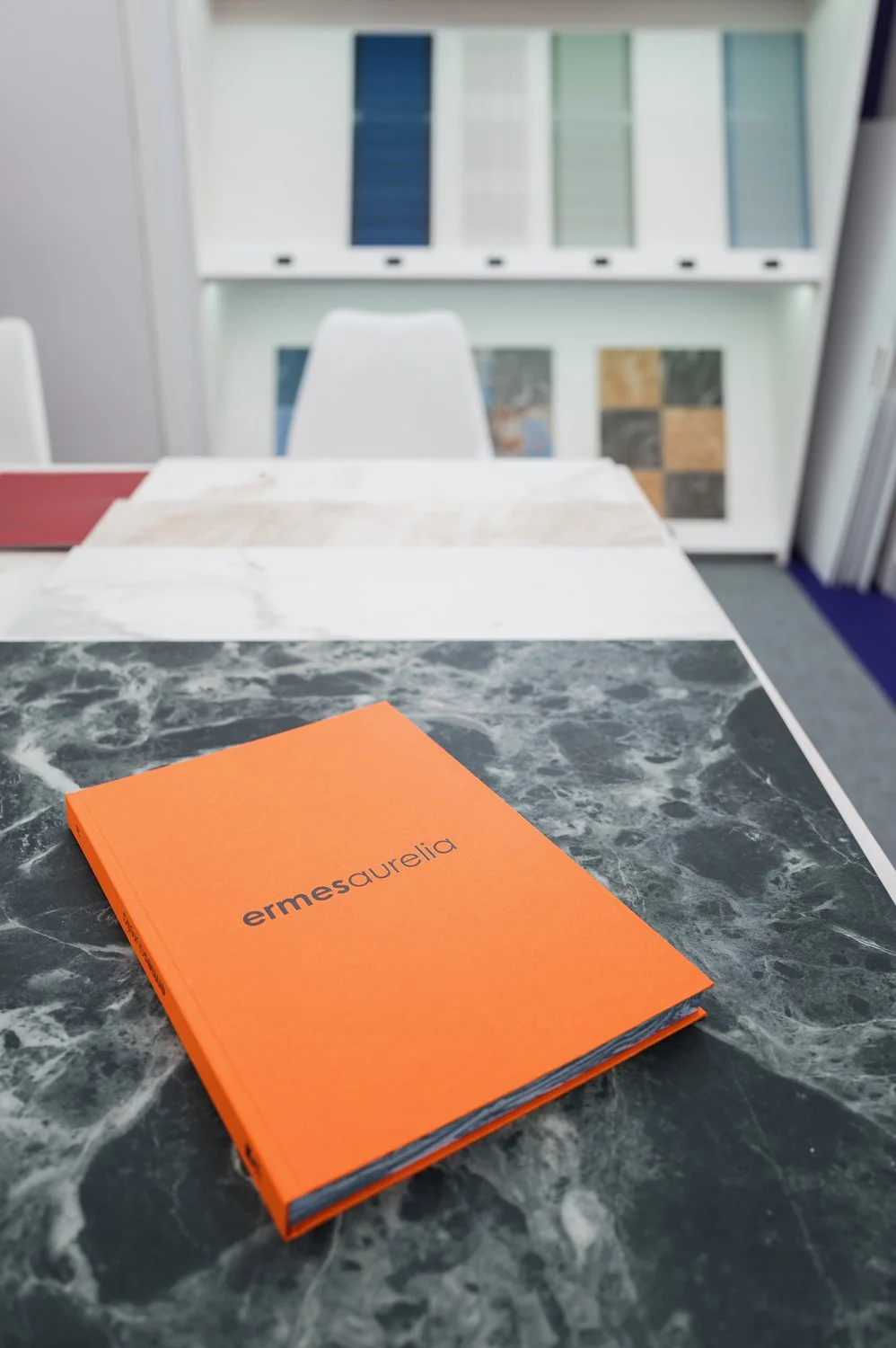 Orange booklet titled 'ermesurelia' on a black marble table in a showroom with fabric samples and tile displays in the background.