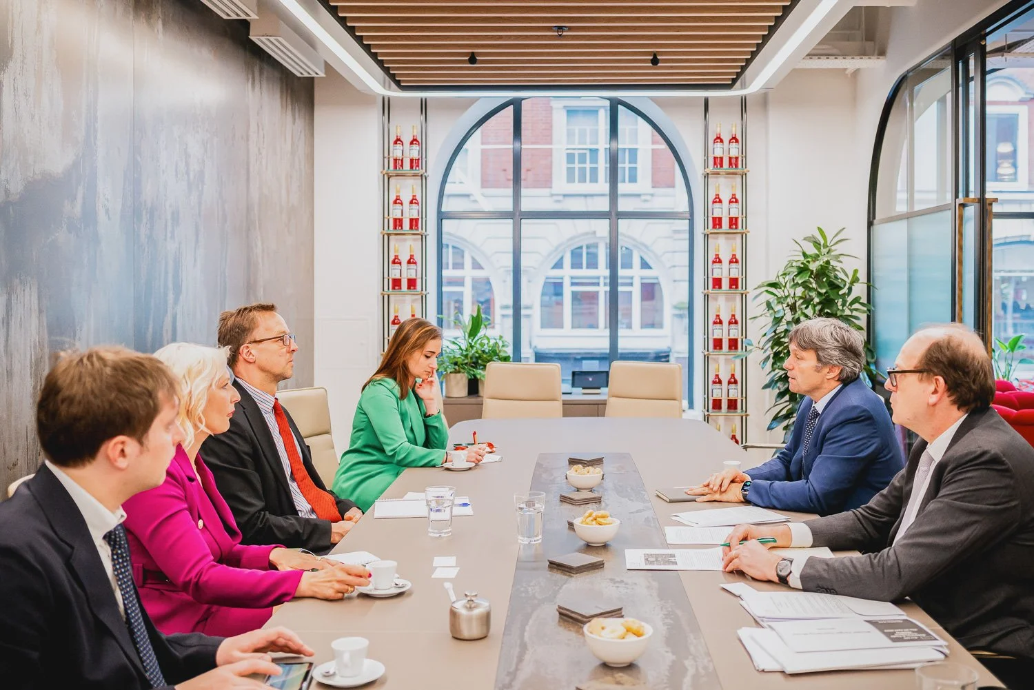 Business meeting with six people seated around a long conference table in a modern room with large arched window and shelves with fire extinguisher bottles.