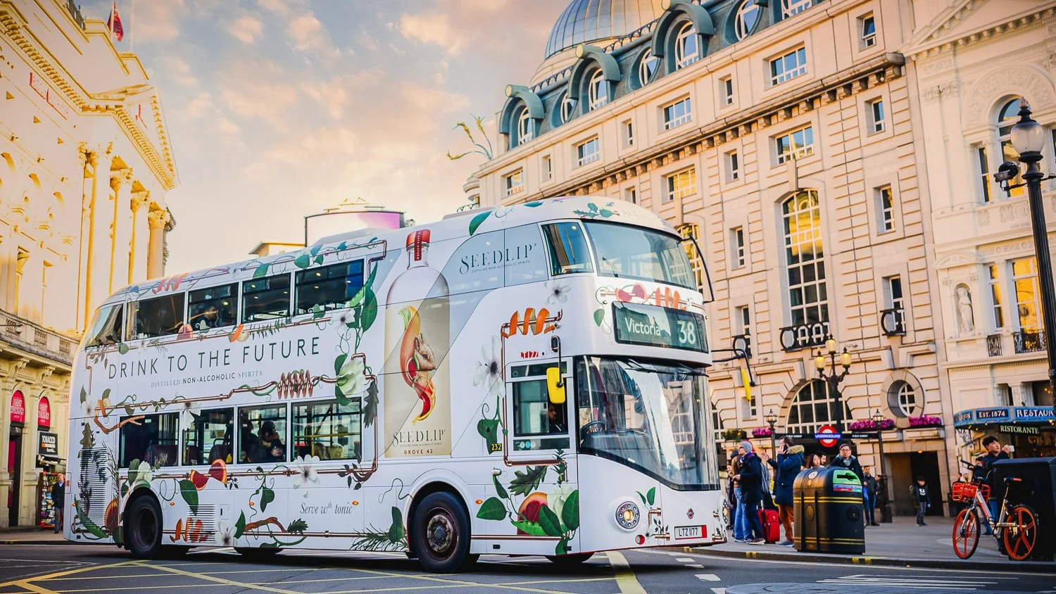 A double-decker bus with colorful food and drink advertisements on city streets, surrounded by pedestrians, bicycles, and historic buildings at sunset. Phocus Collective O-O-H Photography.