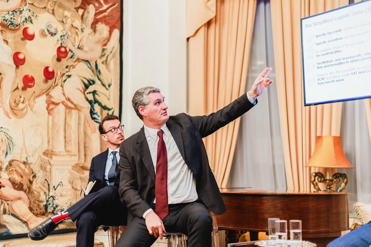 Two men in suits at a presentation or seminar. The man in front is pointing at a screen, while the man behind is sitting and looking up at him. The room has gold curtains and an ornate tapestry on the wall.