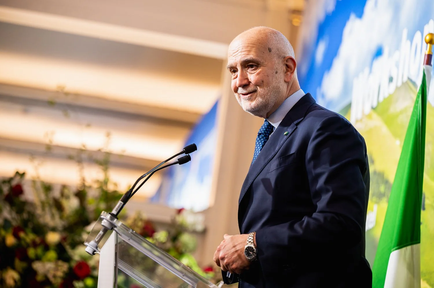 A man in a dark suit and blue tie speaking at a podium, with a microphone, in front of a blue and green backdrop, and an Italian flag to the right.