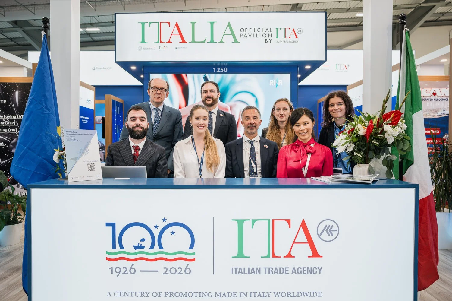 Group of nine people standing and sitting behind a trade booth with a sign reading Italian Trade Agency, celebrating 100 years from 1926 to 2026. The booth has the Italian flag on the right and a blue flag on the left, and a large display screen behi