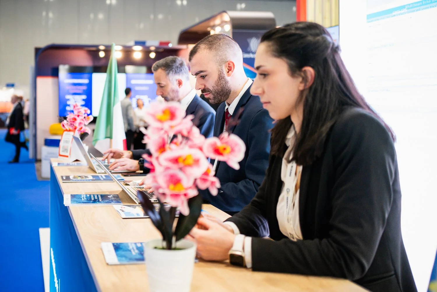 Three people in business attire working at a conference registration desk, with pink flowers and a small Italian flag on the counter.