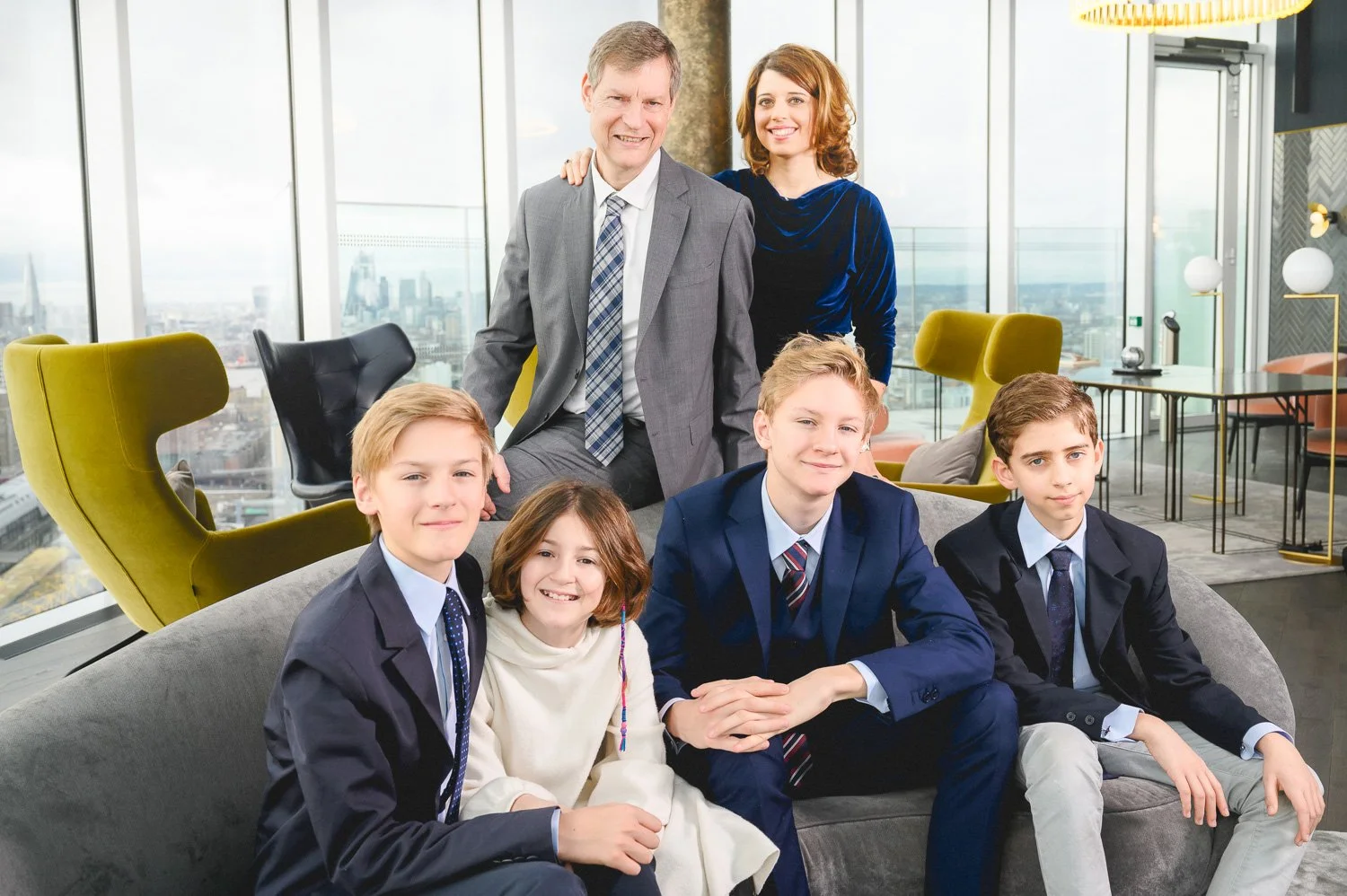A group of three boys and two girls in formal suits and dresses sitting and standing in a modern office with large windows and city view.